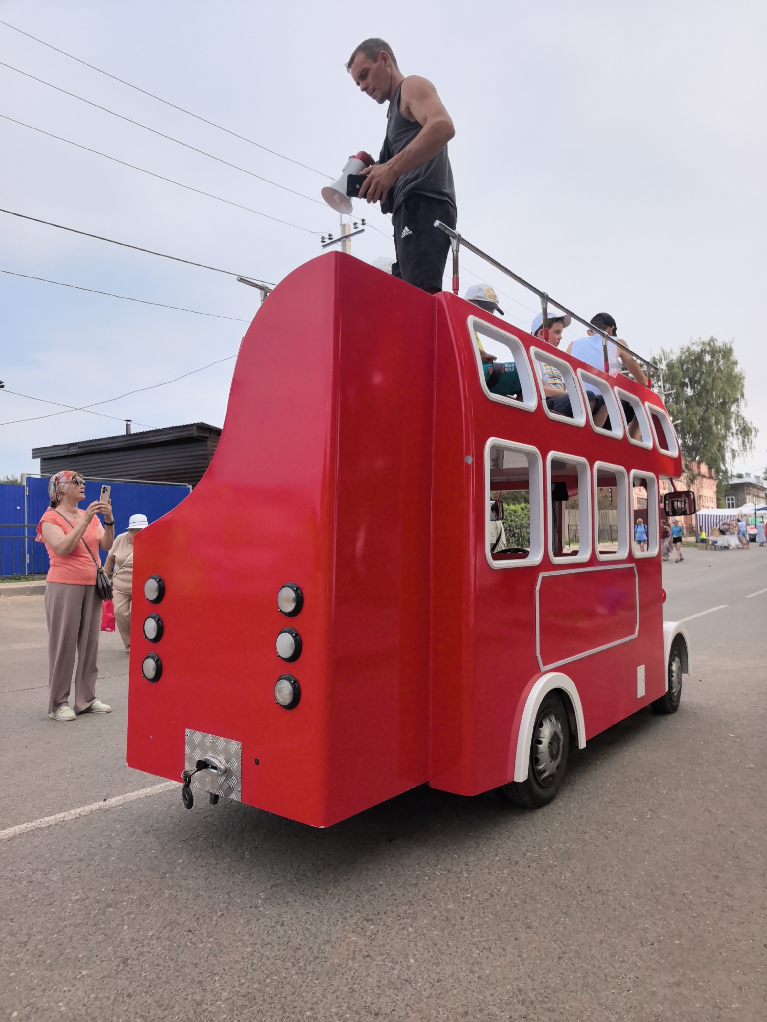 A bright red, two-story bus, resembling a miniature double-decker, is the central subject in the city of Osa, Russia. The bus is parked on an asphalt road, with a white line painted on the left side. The sky is overcast.
On the upper deck of the bus, a man in a grey tank top and black shorts holds a white megaphone and a mobile phone. He is looking down at the phone. Two young boys are also on the upper deck, one wearing a white cap and a striped shirt, the other wearing a dark baseball cap.
On the lower level of the bus, several people are visible through the windows. To the left of the bus, a woman wearing a patterned headscarf, a coral-colored shirt, and light brown pants is taking a photo with her mobile phone. Behind her, another woman in a light-colored hat and outfit is partially visible.
The bus has white trim around its windows and wheel arches. The rear of the bus features a silver hitch mechanism with a black cable attached. Six round, silver lights are arranged in two columns of three on the back of the bus.
In the background, a dark blue fence borders a black building. Further down the street, there are stalls with awnings, people browsing, and buildings, suggesting a public event or market. A tree is visible on the right side of the image.