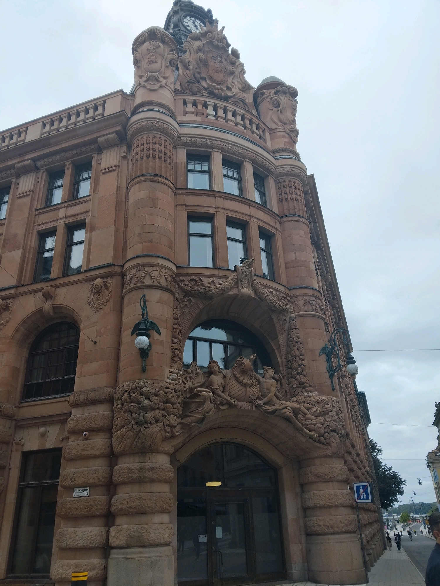 The image shows a street view of the Östermalms Saluhall building in Stockholm, Sweden. The building is made of sandstone and features elaborate architectural details, including ornate carvings, arched windows, and decorative sculptures above the entrance. The top of the building is crowned with a clock tower. The sky is overcast, suggesting a cloudy day. In the foreground, a sidewalk runs alongside the building, with a few pedestrians walking. A street sign labeled "Grevgatan" is visible on the side of the building. A blue pedestrian crossing sign is also present. The street continues into the distance, lined with trees and other buildings.