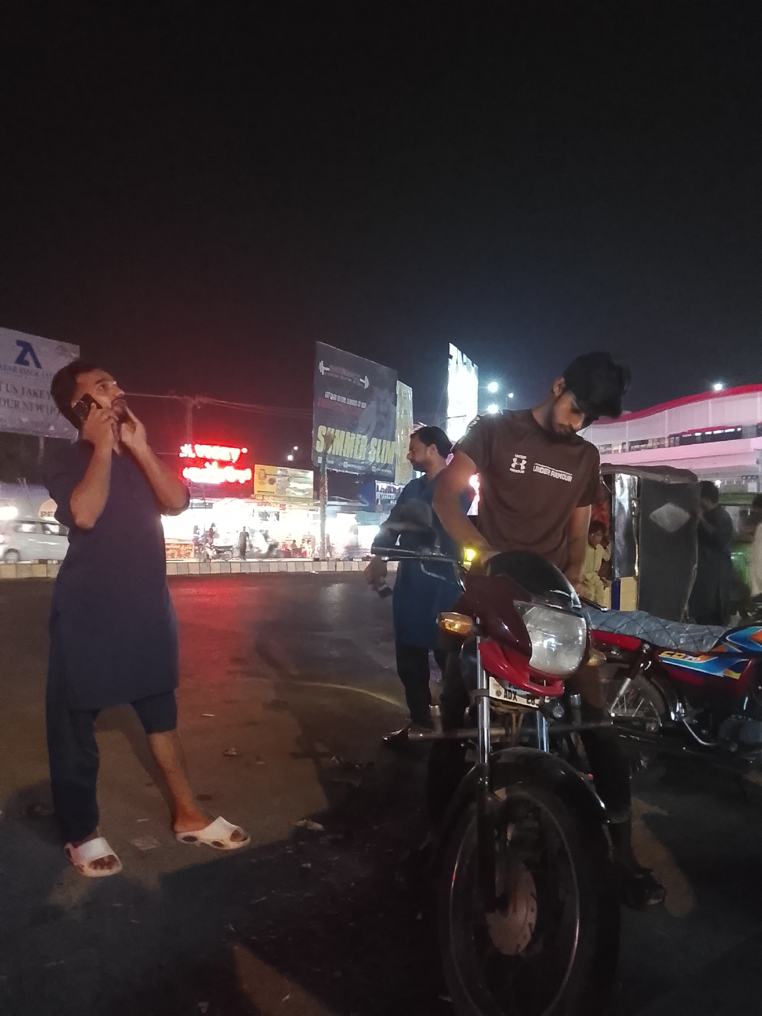 The image captures a nighttime urban street scene, likely a market or commercial area. In the foreground left, a man wearing a dark blue shalwar kameez and white slide sandals is speaking on a mobile phone, looking slightly upwards and to his right. In the foreground right, another man, wearing a brown t-shirt with an "UNDER ARMOUR" logo and a dark cap, stands next to a red motorcycle. He appears to be looking down at the motorcycle or adjusting something on it. The motorcycle's license plate partially shows "ADX 28".
Behind these two individuals, the street extends into a bustling area with various shops and other people, though most are blurred due to motion or distance. A white car is partially visible on the far left, and an auto-rickshaw is visible in the mid-ground right. The scene is illuminated by numerous artificial lights from streetlights and shop signs. Red neon signs, including one with largely illegible text, are prominent in the middle background. Billboards are visible on the left, one partially showing "AEROLINK" and "WE MAKE YOUR DREAMS COME TRUE", and another further right displaying "SUMMER SLIM". The overall atmosphere suggests a typical evening gathering or commute in a busy marketplace.