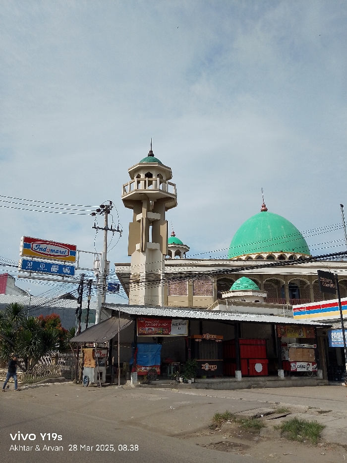 The image shows a mosque in Jatibarang, Indonesia, located at the coordinates -6.471 latitude and 108.302 longitude. The mosque has a tall, thin minaret on the left side and a large, green dome in the center. There is a smaller dome on the right side of the building. The mosque is constructed of white concrete and has a number of arched windows. In front of the mosque, there are a few small shops and food stalls. A person is walking on the sidewalk. The sky is blue and clear, with some clouds in the distance. The ground is paved with gray concrete and there are a few power lines running overhead.