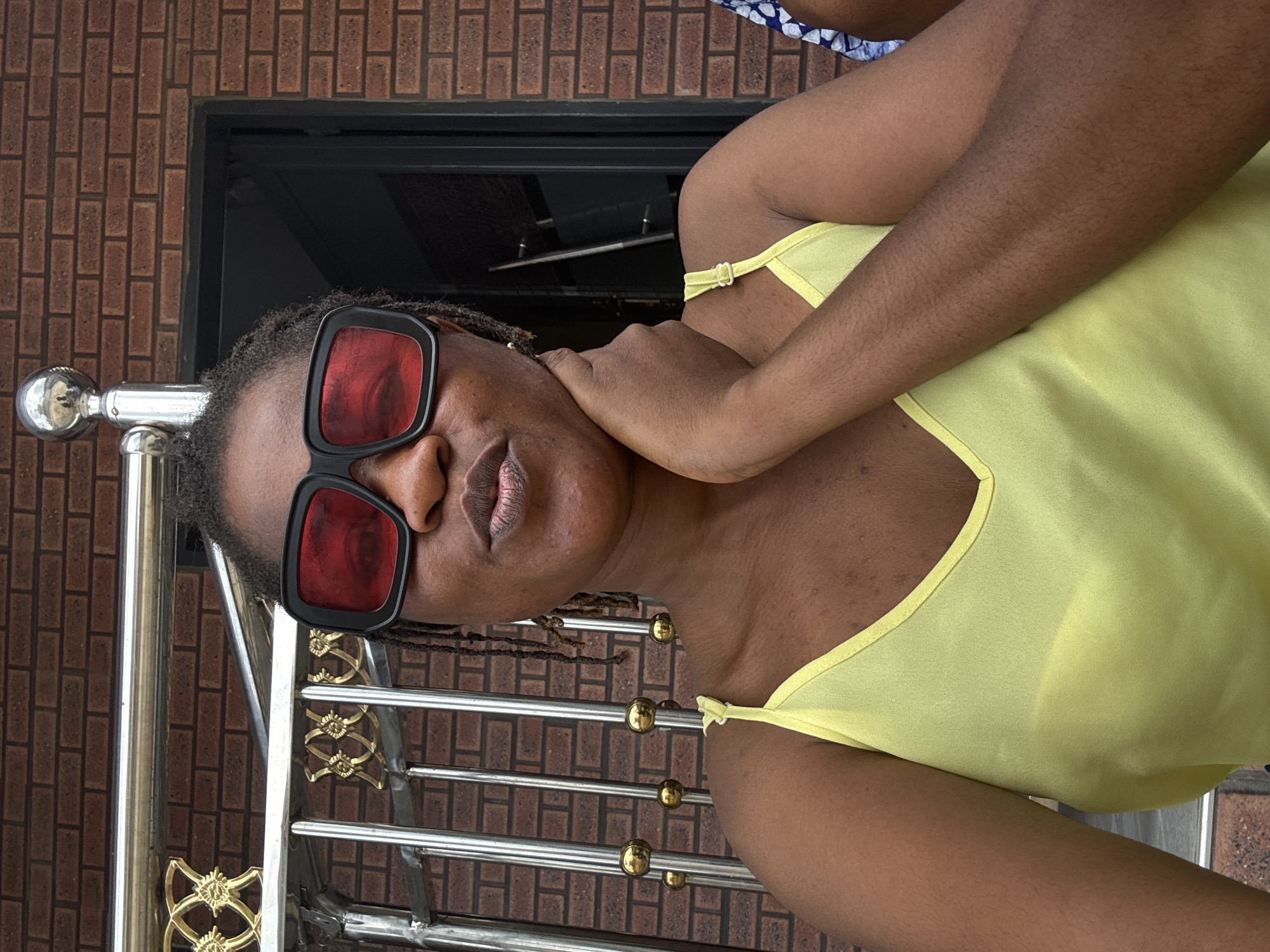 The image shows a selfie of a woman in Ajara, Nigeria. She is standing in front of a brick building with a metal railing. She is wearing a yellow tank top and large black-framed sunglasses with red lenses. Her right hand is resting on her neck, and she is puckering her lips. The railing has ornate gold detailing and is made of chrome. The background shows a brick wall and a doorway.
