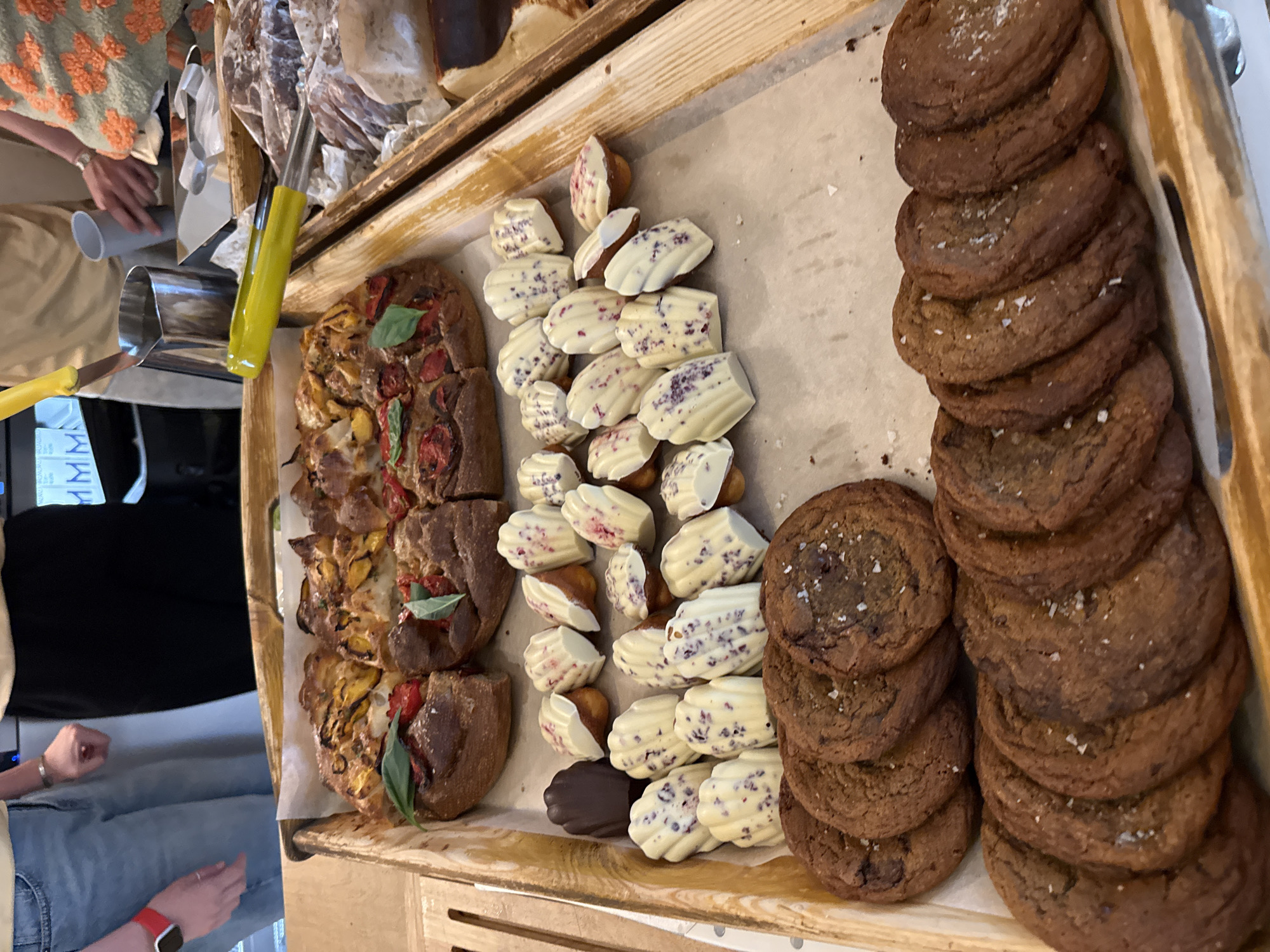 In a bakery or food establishment in Moscow, Russia, a wooden tray displays an assortment of baked goods. The tray is filled with a variety of treats, including several chocolate cookies with visible sea salt crystals, a focaccia-style bread topped with tomatoes and herbs, and a pile of small, shell-shaped cakes, possibly madeleines, which are partly coated in white chocolate with purple speckles. One dark chocolate madeleine is also present.
To the left of the tray, a person's hands are visible. One hand, wearing a silver bracelet, appears to be holding a grey cylindrical object, possibly a container or utensil. This hand is reaching towards the tray. The person is wearing a light-colored shirt and denim jeans. Another hand, wearing a red smartwatch, is also visible, resting on the jeans.
In the background, other food items are partially visible, including what looks like cured meats or pastries in packaging and another tray with more baked goods. A portion of a patterned fabric, possibly clothing or a table runner, is also seen. The setting suggests a retail environment where food is being presented for sale or prepared. The lighting suggests it could be daytime indoors.