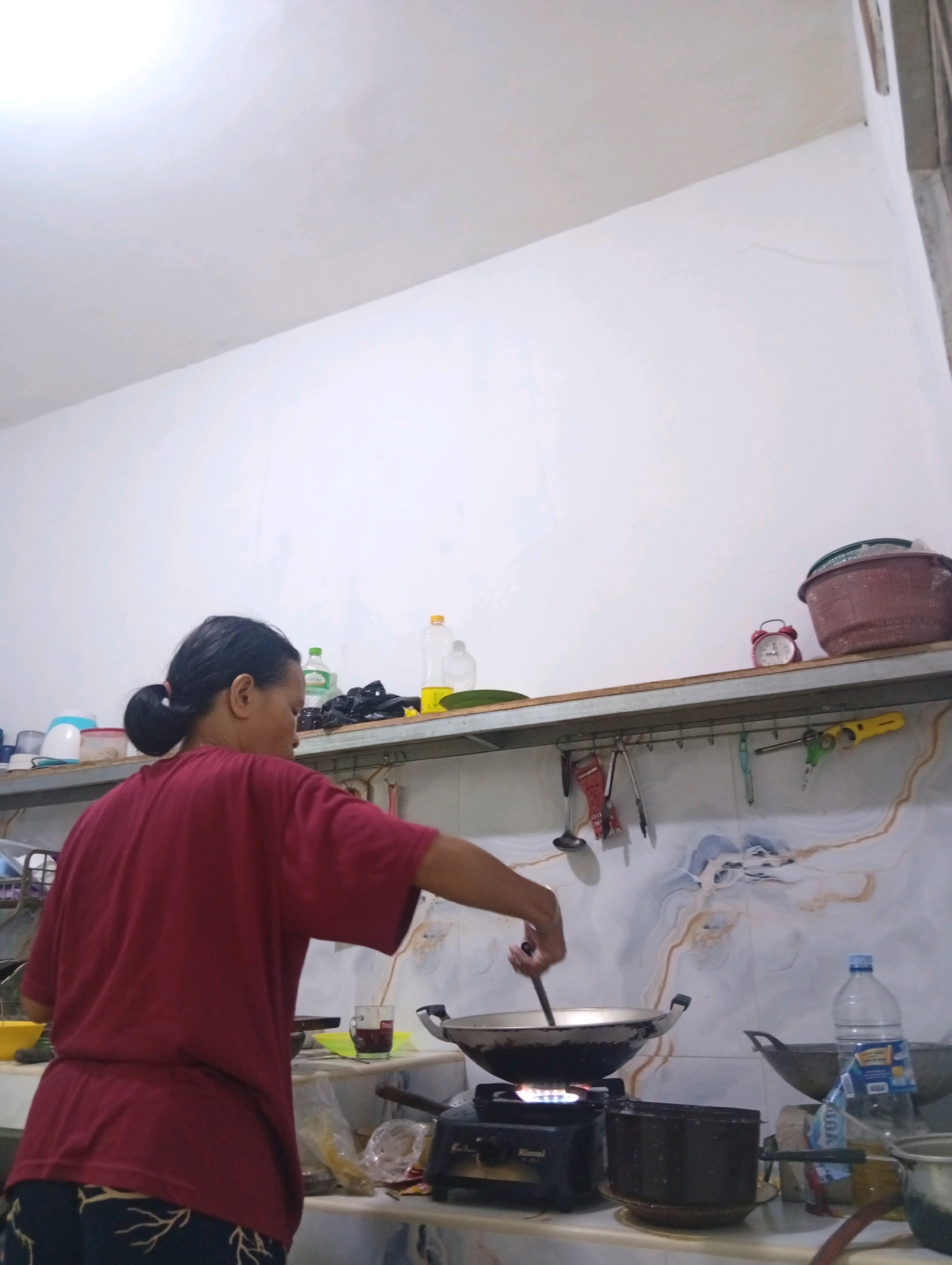 A woman, viewed from the back and right side, with her dark hair tied in a ponytail, is cooking in a kitchen. She is wearing a red t-shirt and dark patterned pants. She is standing in front of a stove with a wok on it, using a utensil to stir the contents. A gas stove is visible below the wok. The kitchen has white walls with some decorative tiling in the background. Shelves above the counter hold various items including bottles, a red bucket, and a red alarm clock. Utensils are hanging from hooks on a shelf below the upper shelves. A bottle of water is visible on the counter next to another pot. The environment appears to be a domestic kitchen in Kebandingan, Indonesia.