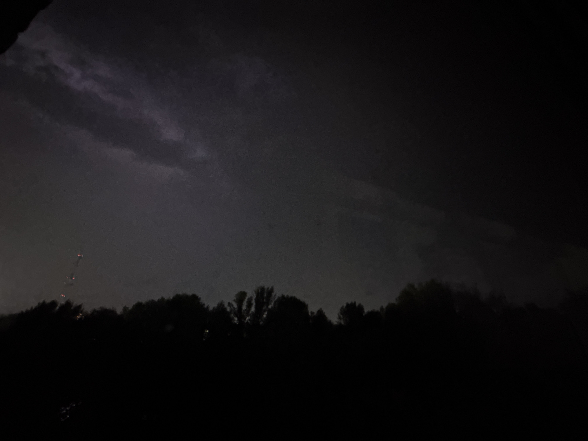 The image depicts a dark, stormy sky with faint lightning flashes visible in the distance, illuminating some clouds. Below the sky, a silhouetted line of trees forms the horizon, with a few faint lights suggesting distant human activity or structures amidst the foliage. The overall scene is one of a nighttime storm, likely occurring in a rural or semi-rural setting given the prominent natural elements and the absence of city lights. The weather appears to be inclement, with dark, heavy clouds and the indication of lightning. The time of day is clearly nighttime, and the mood is somewhat ominous due to the storm. There is no visible text in the image. The location context of Oskemen, Kazakhstan, suggests this could be a natural landscape within that region.