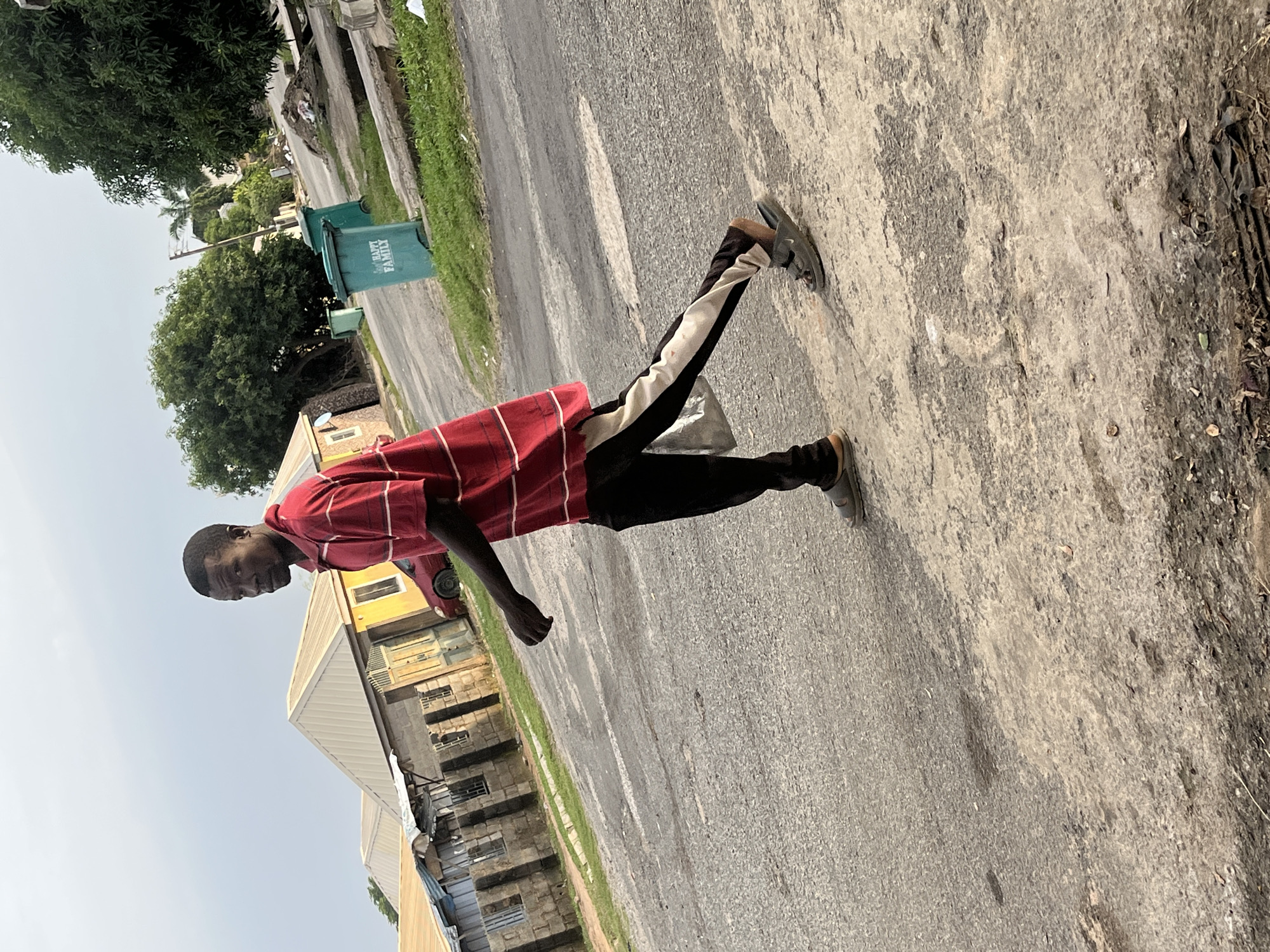 A man walks on a paved road in Abuja, Nigeria. The man is wearing a red and white striped polo shirt, black pants with a white stripe down the side, and sandals. He is carrying a light-colored plastic bag in his left hand. In the background, there are several residential buildings with metal roofs and some trees. Two green trash cans with the label "HAPPY FAMILY" are visible on the right side of the road. The sky is overcast.
