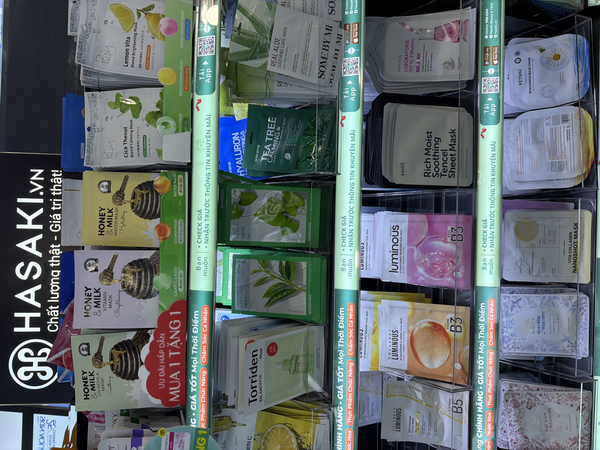The image displays a retail shelf stocked with various face masks. The lighting is bright, suggesting an indoor retail environment. The products are arranged in clear plastic holders, showcasing their packaging. The brands visible include "HONEY & MILK," "Torriden," "INNISFREE," "SOME BY MI," "COLORKEY LUMINOUS," and "DERLABORIE." Some of the masks are promotional, indicated by text like "MUA 1 TANG 1" (Buy 1 Get 1 Free). The shelf also features promotional signage for the "HASAKI.VN" brand and information about downloading their app. The background is dark, likely a wall or display backdrop, with the HASAKI logo prominently displayed at the top. There are no people visible in the image. The environment appears to be a beauty or cosmetic store. The specific location in Bac Nha Trang Ward, Vietnam, is not explicitly depicted, but the presence of Vietnamese text on the signage suggests it is a retail establishment in Vietnam.
