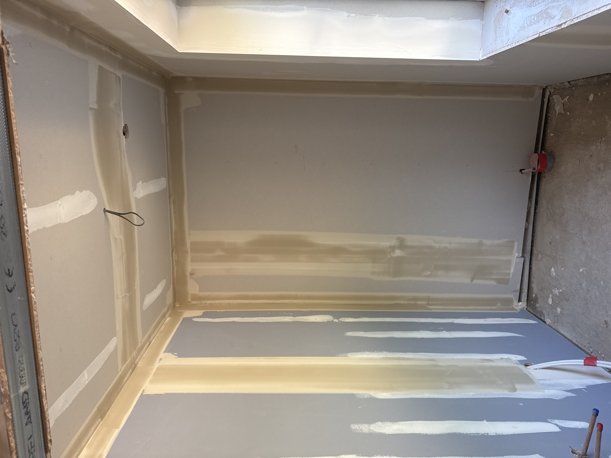 This media file depicts an unfinished interior room, likely a bathroom or utility space, in South Oxfordshire, United Kingdom, undergoing renovation or construction. The walls and floor are covered with plasterboard (drywall), and the surfaces are in the process of being prepared for painting.
Evidence of joint compound application is prominent, with white and beige streaks covering the seams and screw indentations on the plasterboard. On the right wall, a red plumbing fixture with exposed pipes suggests connections for water supply, while additional pipes with red and blue caps are visible on the adjacent floor area. A black electrical wire loop protrudes from a small hole in the left wall. The ceiling features a tiered bulkhead or dropped section, also finished with plasterboard and joint compound.
The scene captures an intermediate stage of interior finishing, specifically the taping and mudding phase of drywall installation. Lighting appears to be artificial. On a metal stud visible along the far left edge, partial text including "CE" and "BS EN" can be seen, indicating compliance with European and British standards, consistent with the specified location. The overall impression is of a functional room being readied for its final finishes.