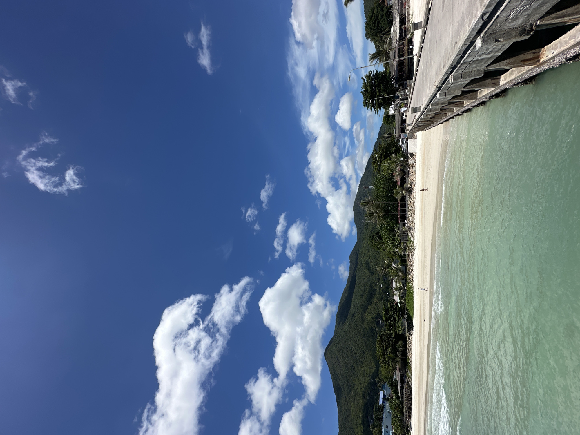The image depicts a bright, sunny day at a beach in Tambon Ko Pha Ngan, Thailand. A long, weathered concrete pier extends into the foreground from the right side of the frame, with its wooden pilings submerged in the clear, turquoise water. Gentle waves lap against a sandy beach in the middle ground, where a few palm trees and low-lying buildings are visible behind a stretch of white sand. In the background, a lush green, tree-covered mountain rises against a vibrant blue sky dotted with fluffy white clouds. A single person is visible walking on the beach, and two other figures are present near the buildings. A white vehicle is parked on the beach near the buildings. The scene captures the serene beauty of a tropical coastal environment.