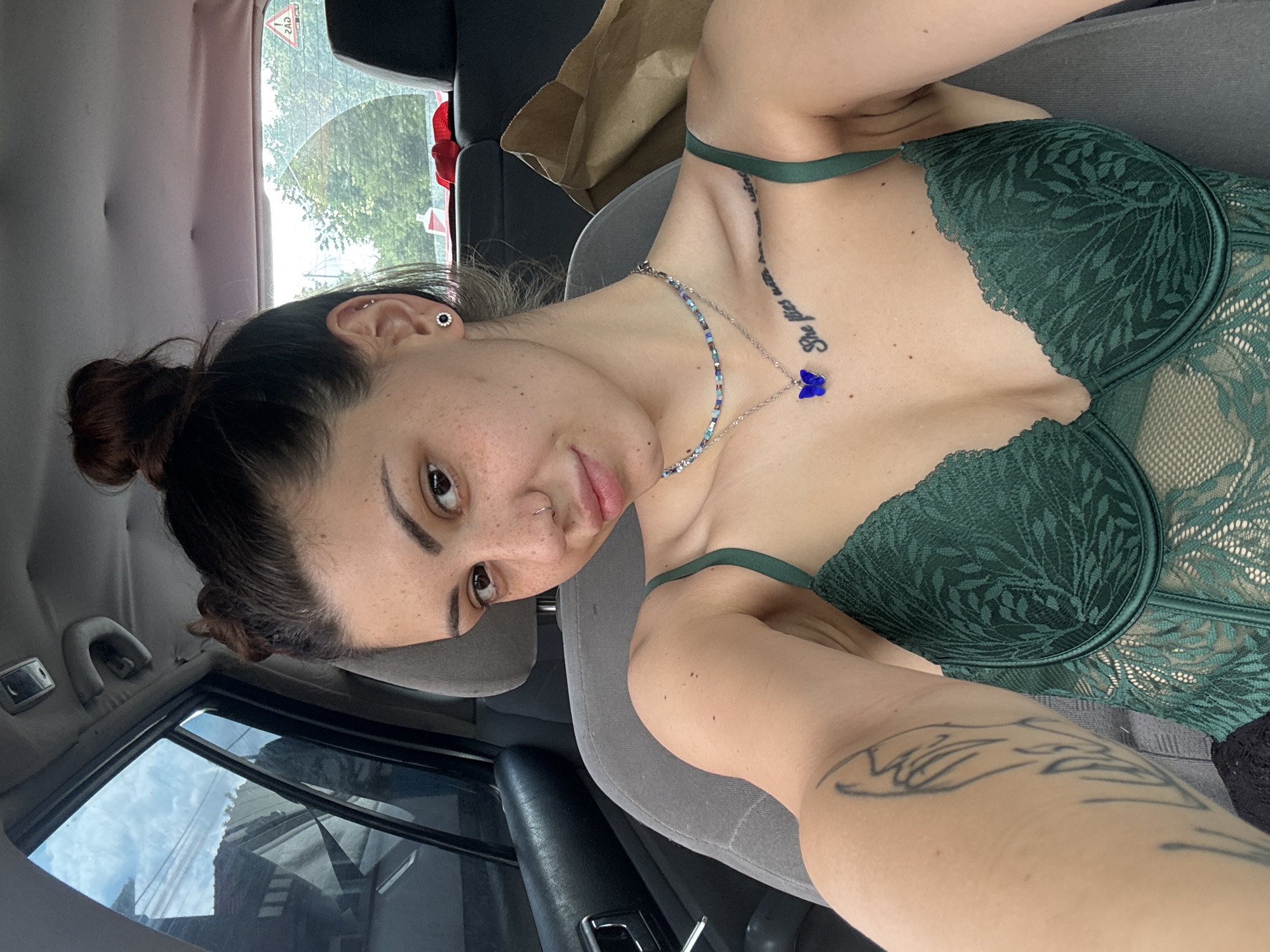 A young woman is captured in a selfie taken inside a car during the daytime. The image is oriented vertically, showing her head tilted to the left and her torso extending to the right, suggesting she is reclining or lying back in a grey fabric car seat. She has dark brown hair styled in a bun, dark eyes, and a scattering of freckles across her nose and cheeks, along with a small silver hoop nose piercing. Her expression is neutral with a subtle hint of a smile.
She is wearing a dark green lace bra or bustier featuring intricate leaf-like patterns. Her accessories include a beaded choker with blue and silver elements, and a silver chain necklace adorned with a distinctive blue butterfly pendant. She also has a stud earring with a dark center and a silver halo, and a small silver hoop in the upper cartilage of her visible ear. Tattoos are visible on her left collarbone, which reads "She flies with" followed by additional text, and a larger, stylized design on her right forearm.
Through the car's rear window, trees and foliage are visible, suggesting an outdoor setting. A reversed red triangular warning sign can be seen attached to the rear window. The side window reflects a cloudy sky and what appears to be urban structures. The environment suggests she is in Gorna Malina, Bulgaria.