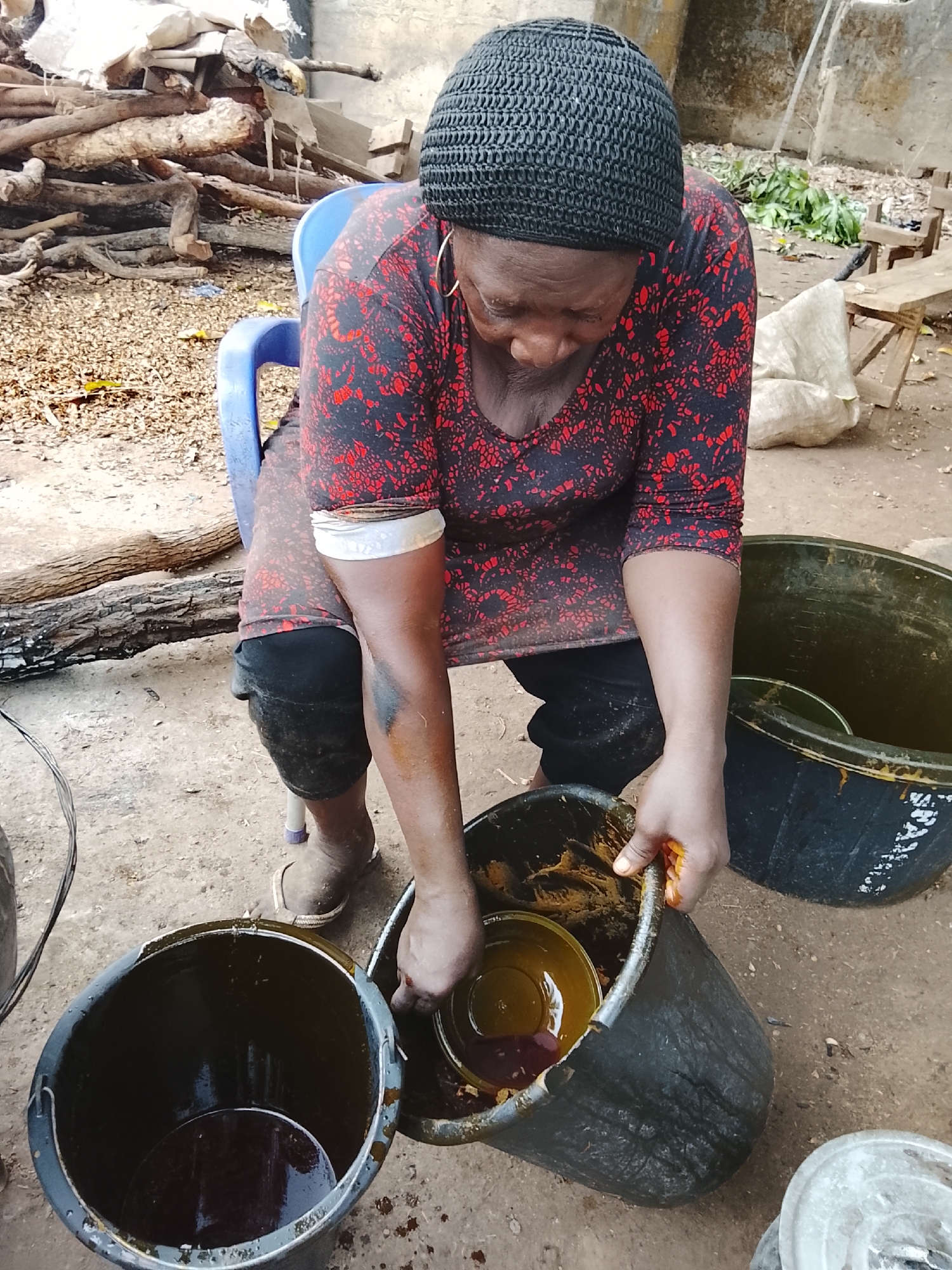 In Akwanga, Nigeria, an elderly Black woman wearing a black crocheted cap and a red and black patterned long-sleeved dress is seated on a blue plastic chair. She is leaning forward, her hands inside a large black plastic bucket containing a yellow liquid, seemingly transferring it into another bucket. A third similar bucket sits to her left, also containing a liquid. The ground appears to be dirt or packed earth. Behind her is a pile of logs, a white sack, and a wooden bench. The setting is likely an outdoor or semi-outdoor space.