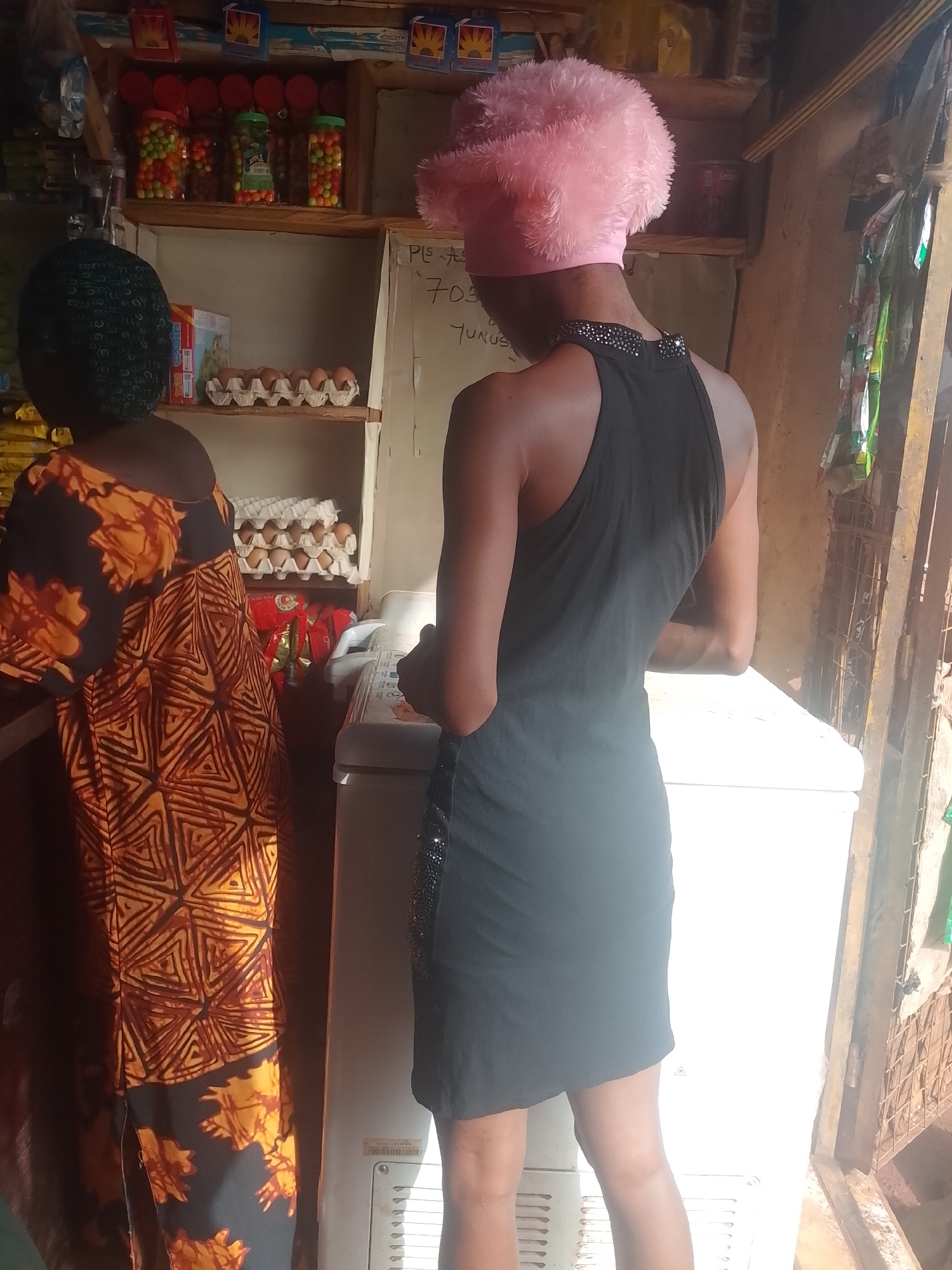 The image shows two individuals inside a shop in Ejule, Nigeria. One person wears a black sleeveless dress and a pink head covering, standing in front of a white refrigerator. The other person, with a dark green head covering, is wearing an orange and black patterned dress and is standing near the counter. Behind them, shelves are stocked with various items, including eggs and jars of multicolored candies. A whiteboard with writing is visible on the wall. The overall environment suggests a small retail establishment.