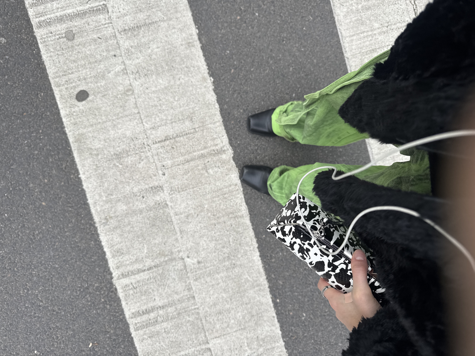 The image shows a street scene, specifically the lower portion of a person standing on a crosswalk. The person is wearing green pants, black shoes, and a black faux fur jacket or coat. They are holding a black and white cow-print purse in their hand. A thin white wire, possibly from headphones or a device, is visible. The background is a crosswalk on an asphalt road. The image captures the subject's feet, the crosswalk stripes, the purse, and the lower part of the garment. The style of clothing and accessories could be indicative of a fashion-conscious individual in an urban environment. Given the architectural style and overall urban aesthetic, it could plausibly be located in a major city, such as Paris, France. No people are visible in the scene other than the lower portion of the person standing on the crosswalk.