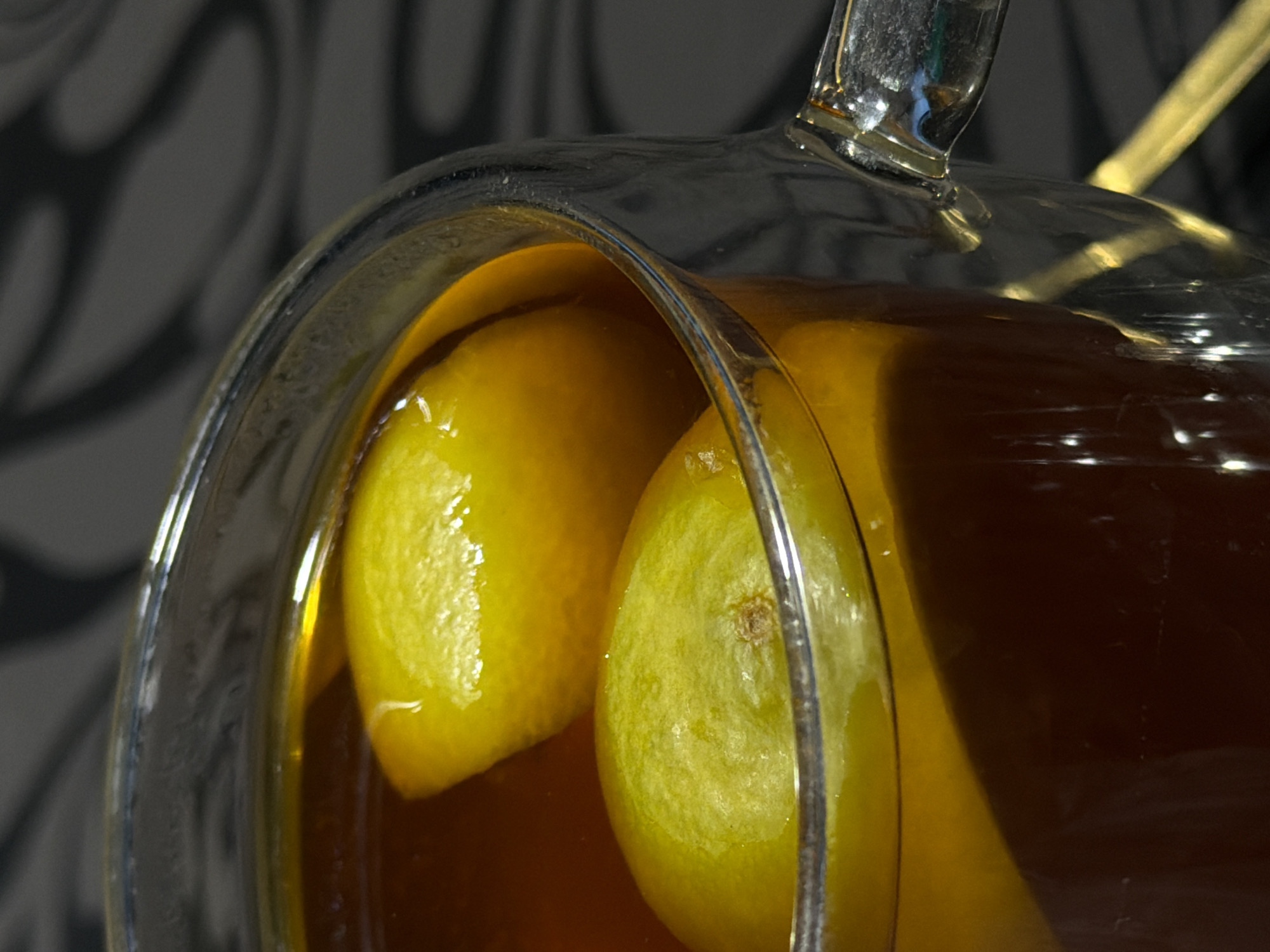 A close-up shot shows a clear glass pitcher filled with a dark amber liquid and two yellow lemon halves. The pitcher has a visible handle. The background features a dark gray wall with indistinct patterns. The image appears to be taken indoors. The location is Rossiyskiy, Russia.