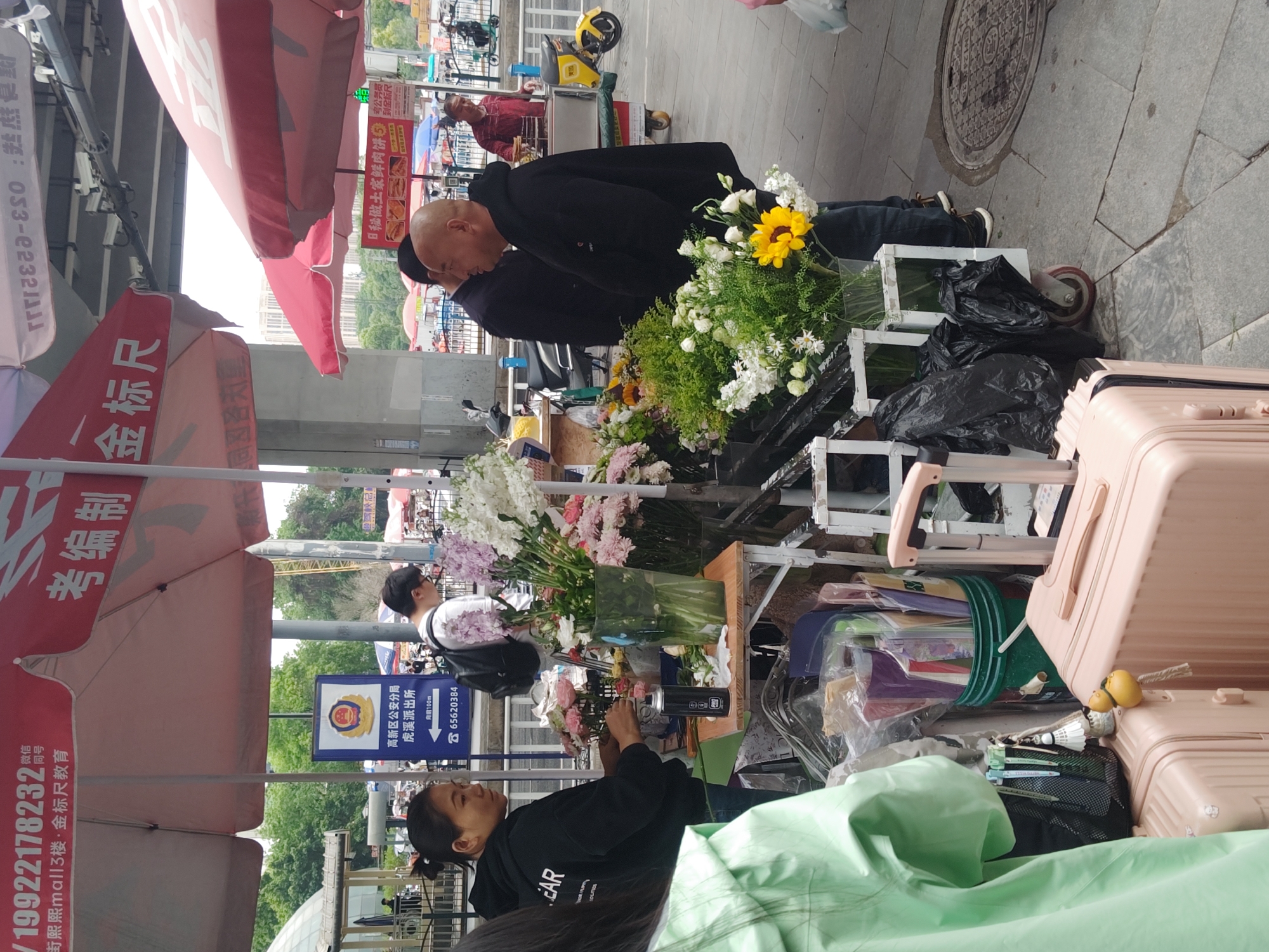 The image shows a street scene in the High-tech Zone of Chongqing, China. A flower stall is set up, displaying a variety of flowers including white lilies, pink carnations, yellow sunflowers, and other mixed bouquets. Two bald men in black jackets are examining the flowers. A woman in a black sweatshirt is arranging flowers. Behind the woman, there is a blue sign that reads: "High-tech Zone Public Security Bureau, Huxi Police Station, 100m ahead, 65620384". A red umbrella with white Chinese characters is above her. A second red umbrella is located behind the two bald men in black jackets. Next to the stall is a pink suitcase. The scene is on a sidewalk with a manhole cover in the background.