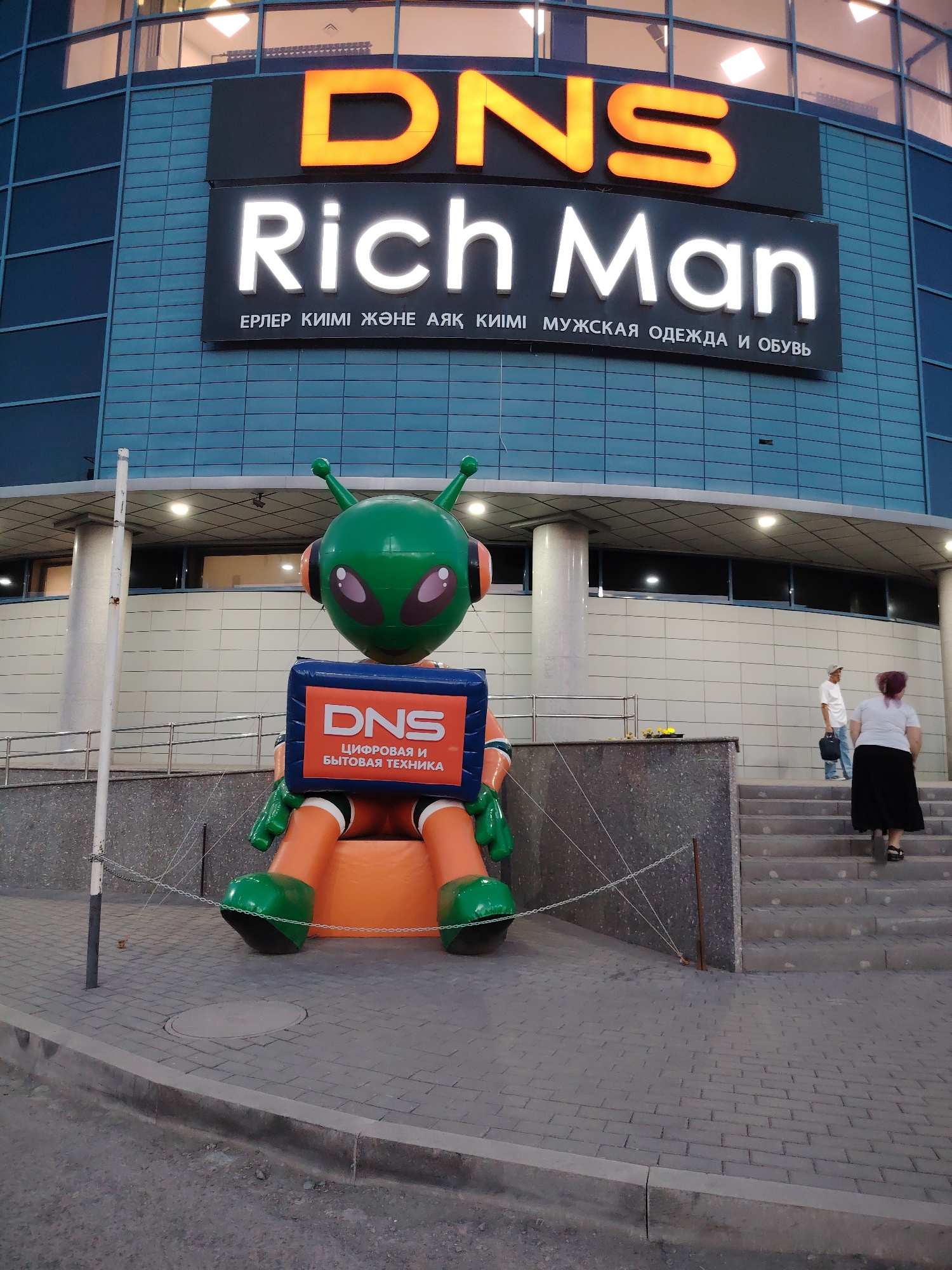 This is a photograph taken in Temirtau, Kazakhstan, showing the exterior of a "DNS Rich Man" store. The store's facade is a prominent blue tiled wall with large illuminated signs. The top sign displays "DNS" in orange, followed by "Rich Man" in white. Below these are two lines of text in what appears to be Cyrillic and possibly Kazakh, with the latter part reading "МУЖСКАЯ ОДЕЖДА И ОБУВЬ," translating to "Men's Clothing and Footwear."
In the foreground, a large, inflatable, cartoonish alien-like robot stands prominently. This robot is green with orange limbs and has a blue chest piece displaying the "DNS" logo and the text "ЦИФРОВАЯ И БЫТОВАЯ ТЕХНИКА," meaning "Digital and Home Appliances." The robot is positioned on an orange base, with its legs and feet visible.
To the right of the robot, a set of concrete stairs leads up to the store entrance. Two people are visible on the stairs: a man in a white shirt and a cap carrying a dark bag, and a woman with dark hair wearing a white top and a long black skirt. They are ascending the stairs, suggesting they are entering or leaving the store.
The scene appears to be set in the late afternoon or early evening, as the lighting on the building and the signs indicates the presence of artificial illumination. The pavement in front of the robot is made of cobblestones, and metal chains with posts delineate the area. The overall impression is that of a commercial street or entrance area of a shopping establishment.