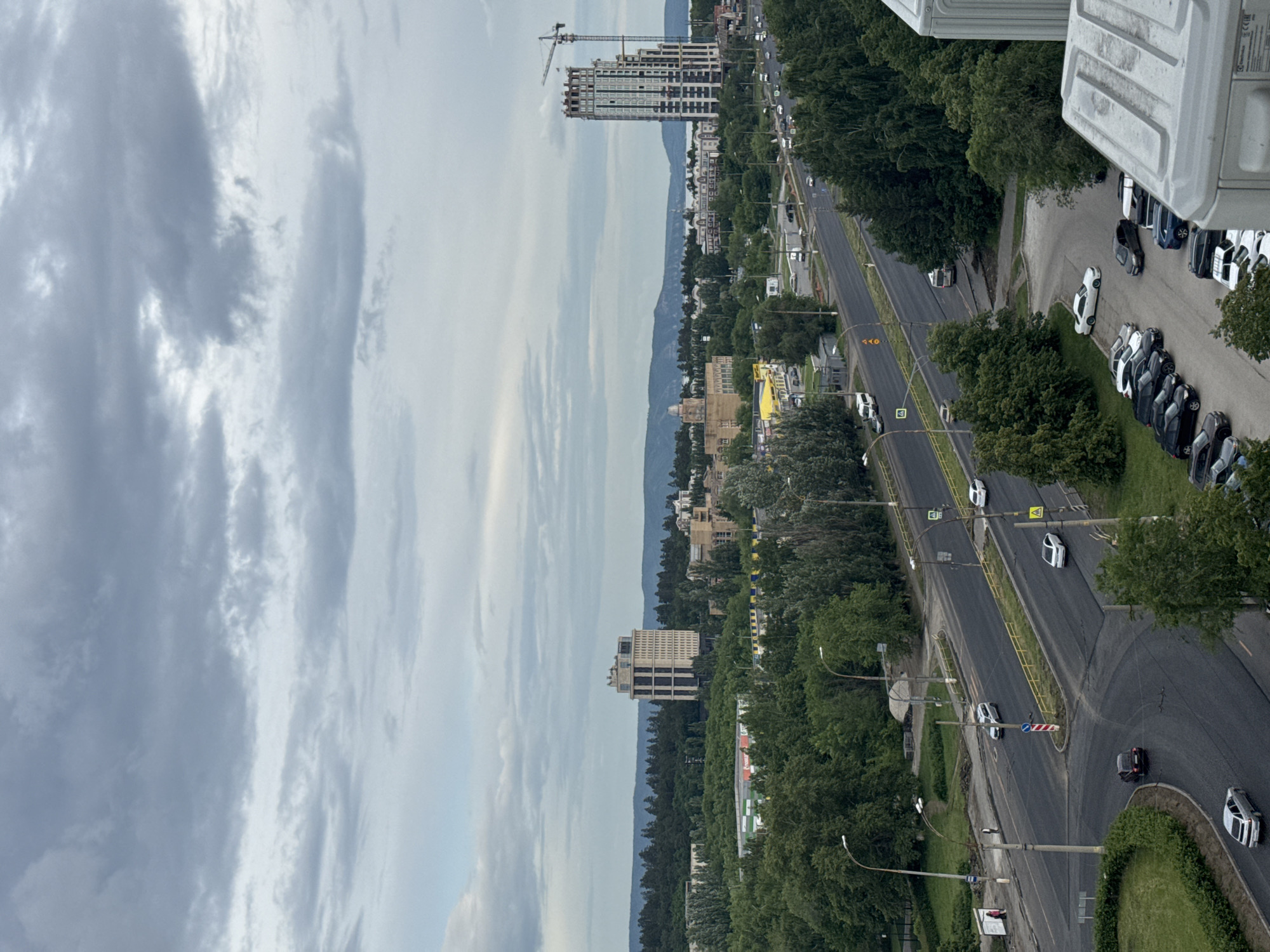 This elevated, wide-angle shot captures a daytime urban landscape under an overcast sky, located in Volgograd, Russia. The scene is dominated by a multi-lane asphalt road, running diagonally through the midground, with various vehicles in motion and parked. Abundant green foliage, consisting of numerous trees and bushes, lines the roads and forms large green spaces, creating a significant natural buffer throughout the city.
In the foreground, partially visible on the bottom right, is the top portion of a light-colored outdoor air conditioning unit or similar utility structure. Below it and to its left, a paved parking lot contains several parked cars of various models and colors, including white, black, and grey sedans.
The main road features yellow solid and dashed lines, as well as white dashed lines, indicating lanes and road edges. Streetlights with double luminaires are spaced along the roadside. Several road signs are visible, including a red circular sign with a white horizontal bar, pedestrian crossing signs, and "A" in a triangle warning signs. On the left side of the road, a silver sedan is visible making a turn at a small, rounded intersection, while a black sedan is further down, also turning. Several other vehicles, including a white van, are visible on the road and at the edges. A bus stop shelter with a person standing nearby is on the left side of the road.
In the mid-distance, a diverse array of buildings is interspersed among the trees. On the left, a tall, multi-story building with a light beige facade and distinctive dark vertical sections stands prominently. Further to its right, a building with a domed roof is visible, identified as the Palace of Culture named after S. M. Kirov. Nearby, a yellow building with a red sign, partially obscured but showing "MAGNUM", suggests a commercial establishment.
To the right, a very tall residential building, known as "Volga Towers," is under active construction, indicated by the presence of a large tower crane positioned next to it. This building features a modern, stepped design with visible green architectural elements. Beyond the buildings and the dense tree line, a wide body of water, likely the Volga River, is visible, reflecting the overcast sky. In the far background, a range of low hills or mountains stretches horizontally across the horizon under the grey clouds.