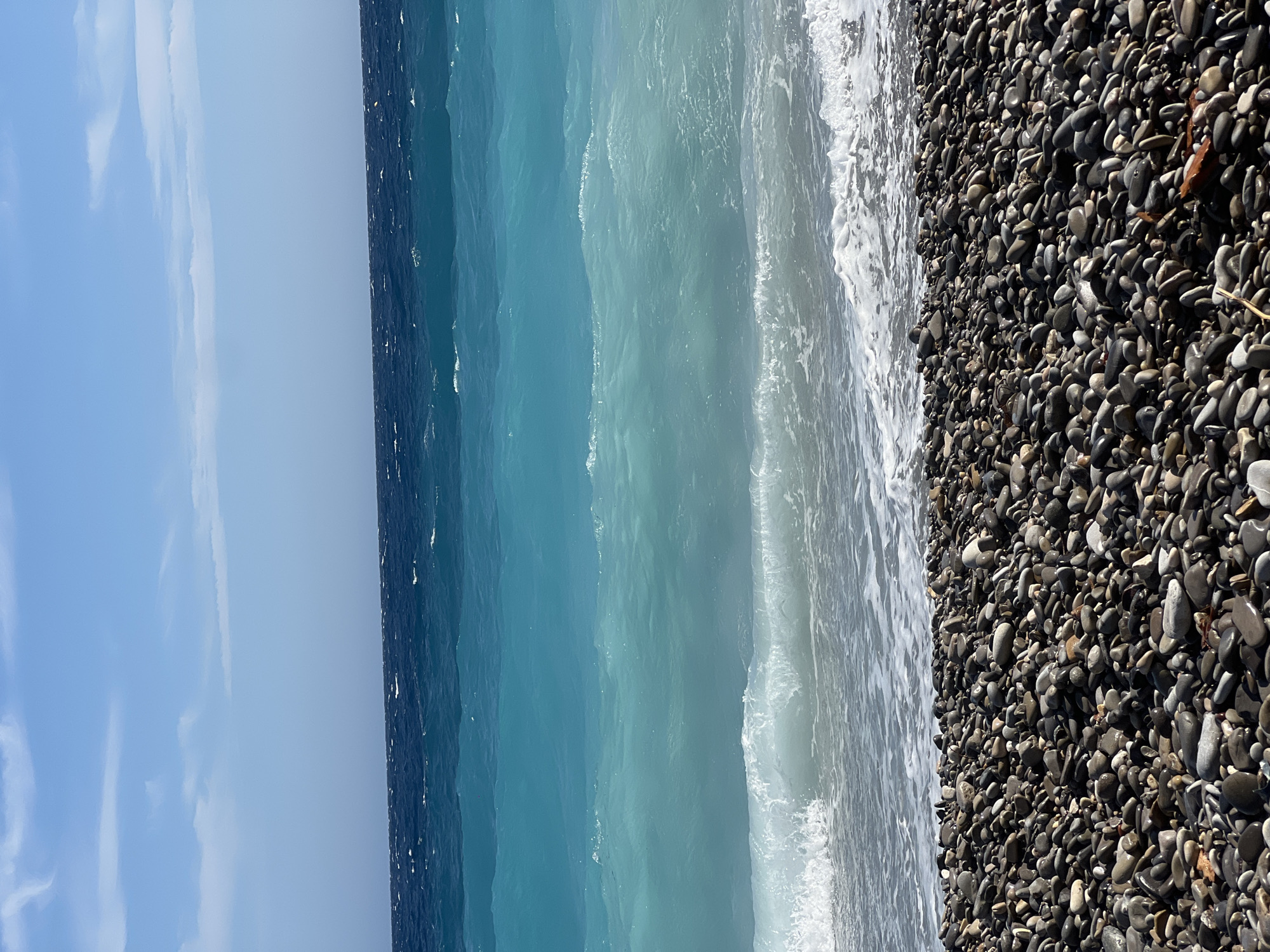 A beach in Nice, France, is depicted with a foreground of dark, smooth pebbles. Beyond the beach, the Mediterranean Sea exhibits distinct bands of color, transitioning from a bright turquoise near the shore to a deep navy blue further out. Gentle waves are breaking on the pebbled shore, creating white foam. The sky above is a pale blue with scattered white clouds. No people are visible in the image.