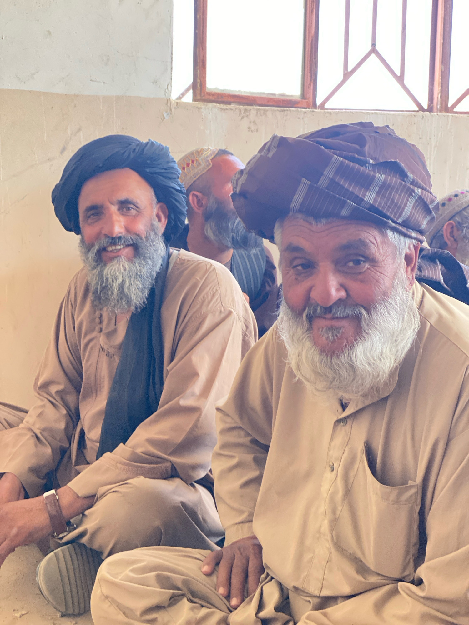 A medium shot captures three adult males, seated in a row indoors. The two men in the foreground, with their faces turned towards the viewer, are the primary subjects. Both men are wearing traditional clothing consisting of loose-fitting tunics and large turbans. The man on the left has a dark blue turban and a grey beard, while the man on the right wears a brown and white striped turban and a white beard. Their expressions appear relaxed and friendly. The background is a plain, textured wall with a window at the top. The third man is partially visible behind the man on the left, also wearing a turban. The lighting suggests a bright, possibly outdoor, environment that is being filtered through the window. The setting appears to be a simple, rustic interior space, potentially a community building or a home. The floor is covered in what appears to be sand or dirt. This image was captured in Pasab, Afghanistan.
