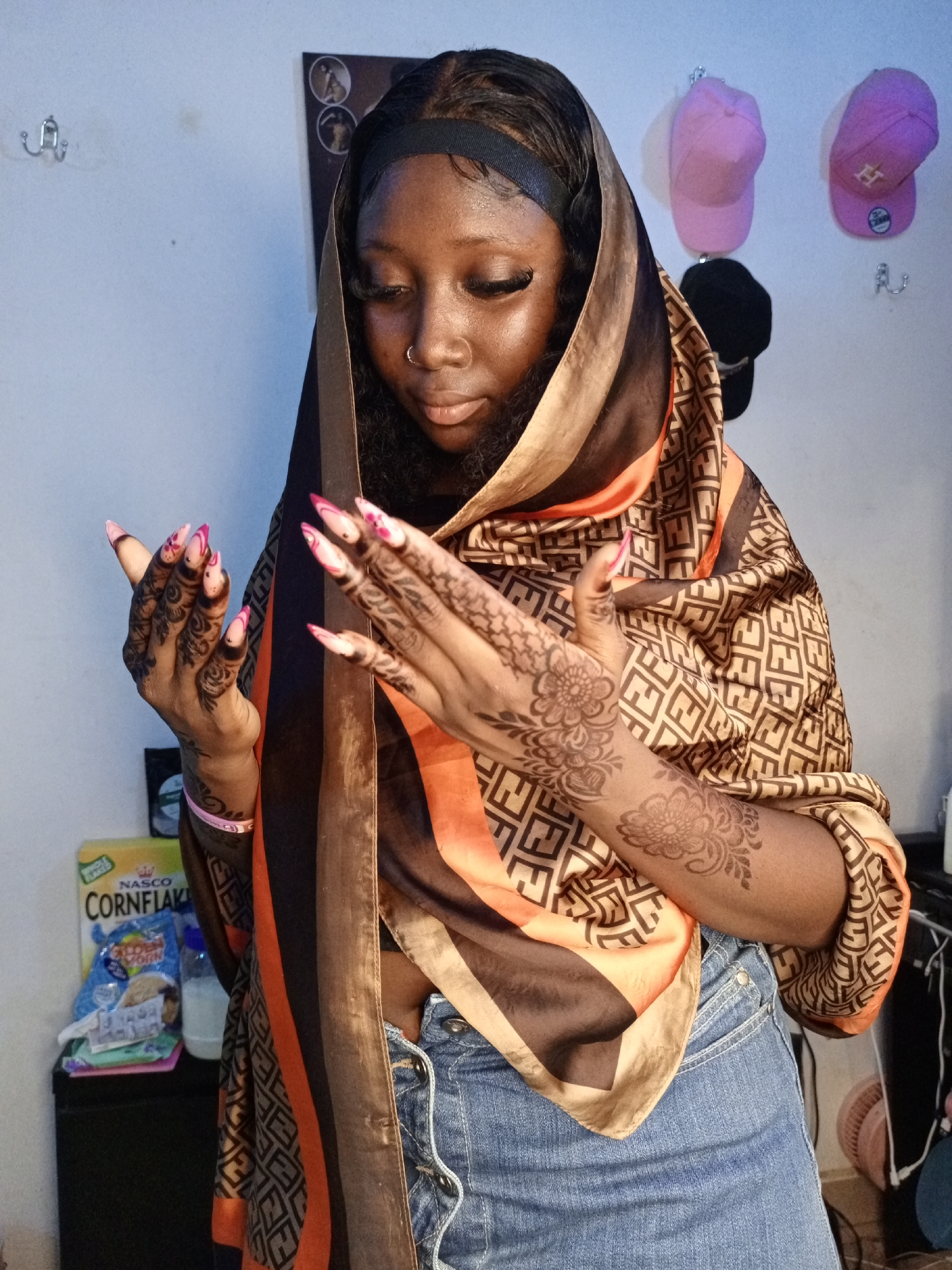 A woman is standing indoors. She is wearing a printed brown and orange scarf over her head and shoulders, a black headband, and blue jeans. She has long, pink-tipped nails and henna tattoos on her hands and arms. The woman has a nose ring and is looking down at her hands. In the background, there is a white wall with three caps hanging on hooks. A box of Nasco Cornflakes and other items are on a small black shelf to the left. The location is Unknown, Unknown.