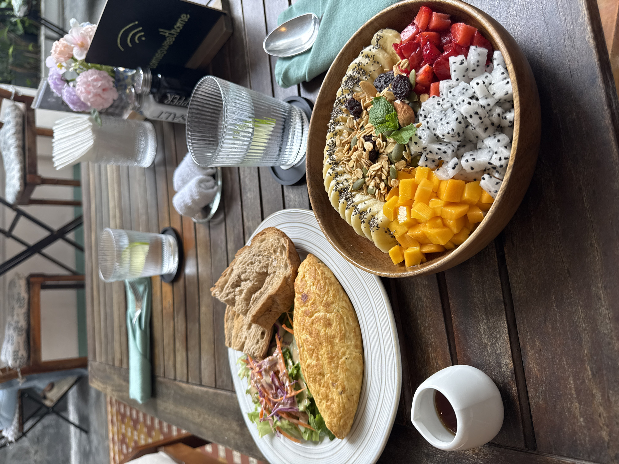 On a dark, wooden table, a meal is laid out. A wooden bowl filled with various fruits including banana slices topped with chia seeds, strawberries, diced mangoes, and pieces of dragon fruit is positioned towards the right. Adjacent to the fruit bowl is a white plate containing an omelet, two slices of bread, and a salad. A small, white container holding a dark liquid sits near the plate. Two glasses of water are present, along with folded napkins and utensils. A book and a vase of flowers are in the background. The setting appears to be a restaurant or dining area, possibly in Đà Nẵng, Vietnam.