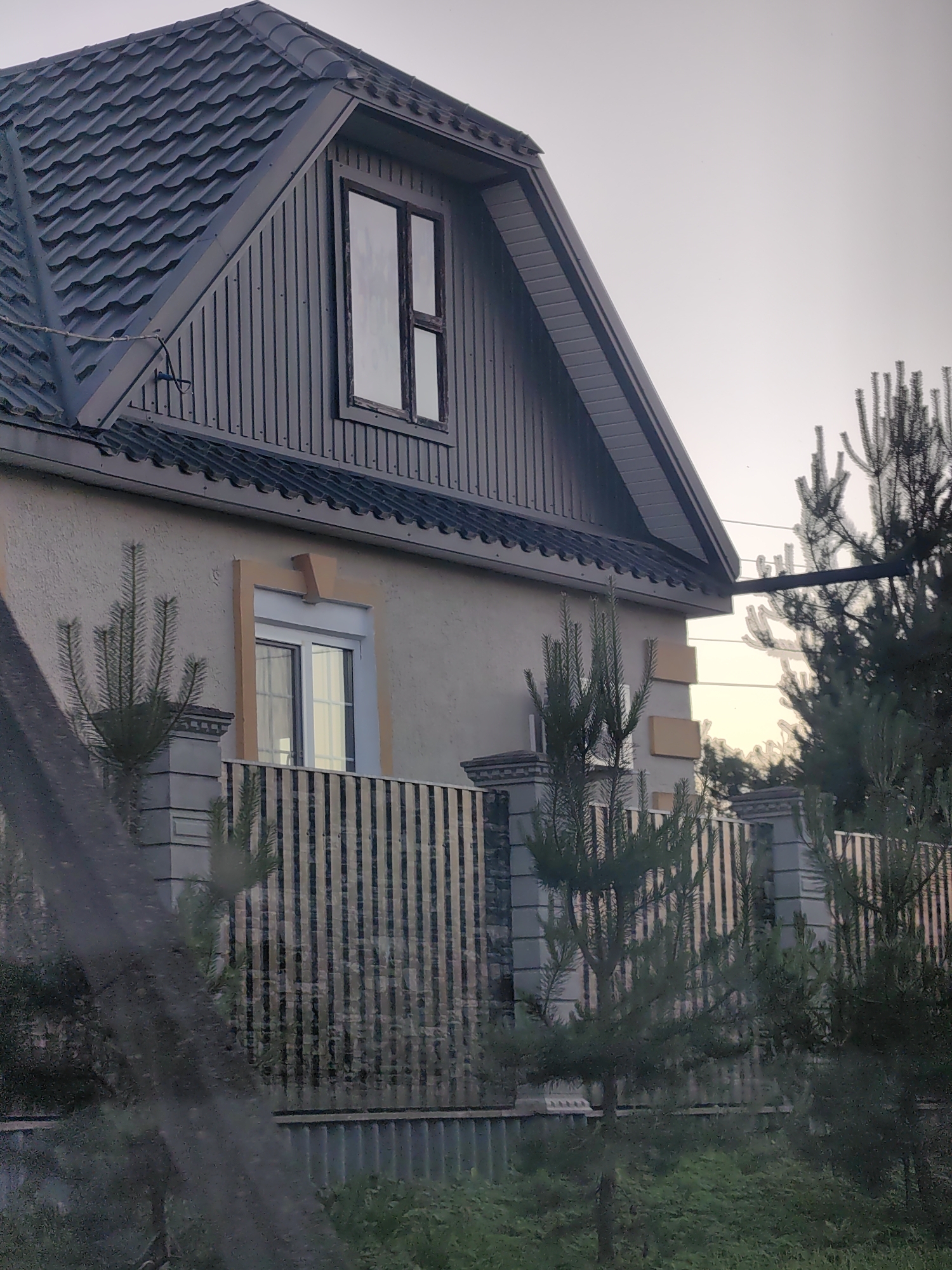 The image shows the exterior of a two-story house with a dark gray metal roof and beige siding. A prominent feature is the steeply pitched roof with visible horizontal ridges. A dormer window with a dark frame is centrally located on the upper story, set into vertical siding. On the lower level, another window with a light-colored frame and an orange decorative surround is visible. A decorative fence runs along the front of the property, constructed with stone pillars and vertical black slats. Small evergreen trees are planted in front of the fence. The surrounding environment appears to be residential, with a clear, overcast sky. The scene is captured from a slightly low angle, suggesting it might be taken from across a street or pathway.