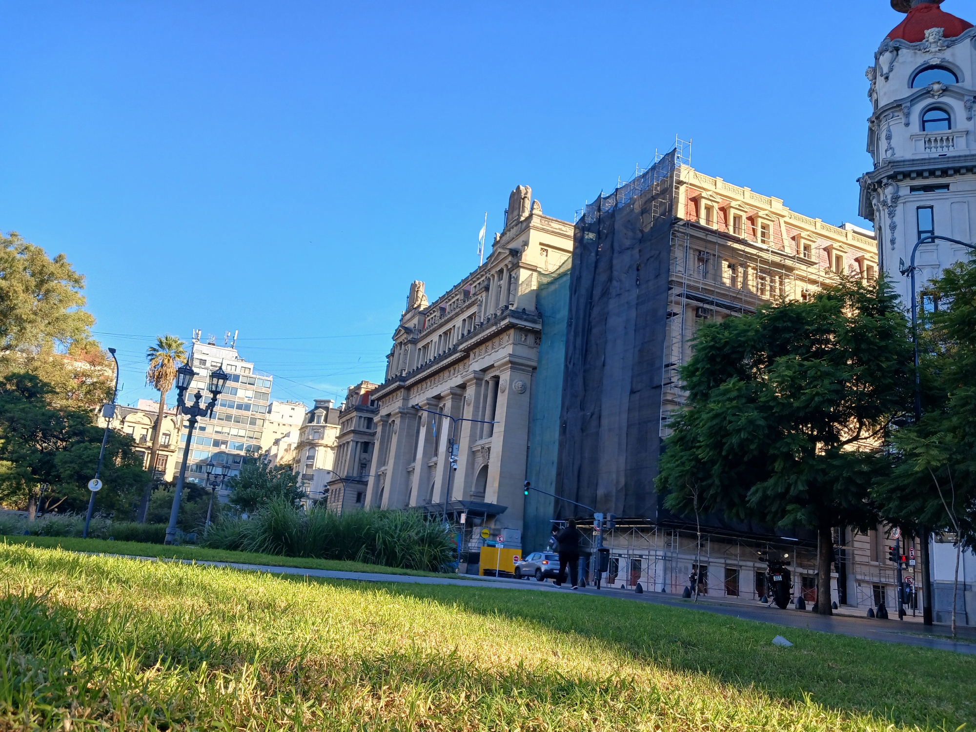 The image shows a low-angle view of a street scene in Buenos Aires, Argentina, featuring government buildings and lush green grass. The most prominent building is a large, ornate structure with classical architectural elements like columns and statues. A portion of the building is covered in scaffolding and dark blue netting, indicating ongoing construction or renovation. A second building on the right side with red dome. A street runs between the grass and the building, with a few pedestrians visible. Cars and traffic lights are present, indicating an active urban environment. The sky is clear and blue, suggesting a sunny day. Several buildings are in the background, to the left.