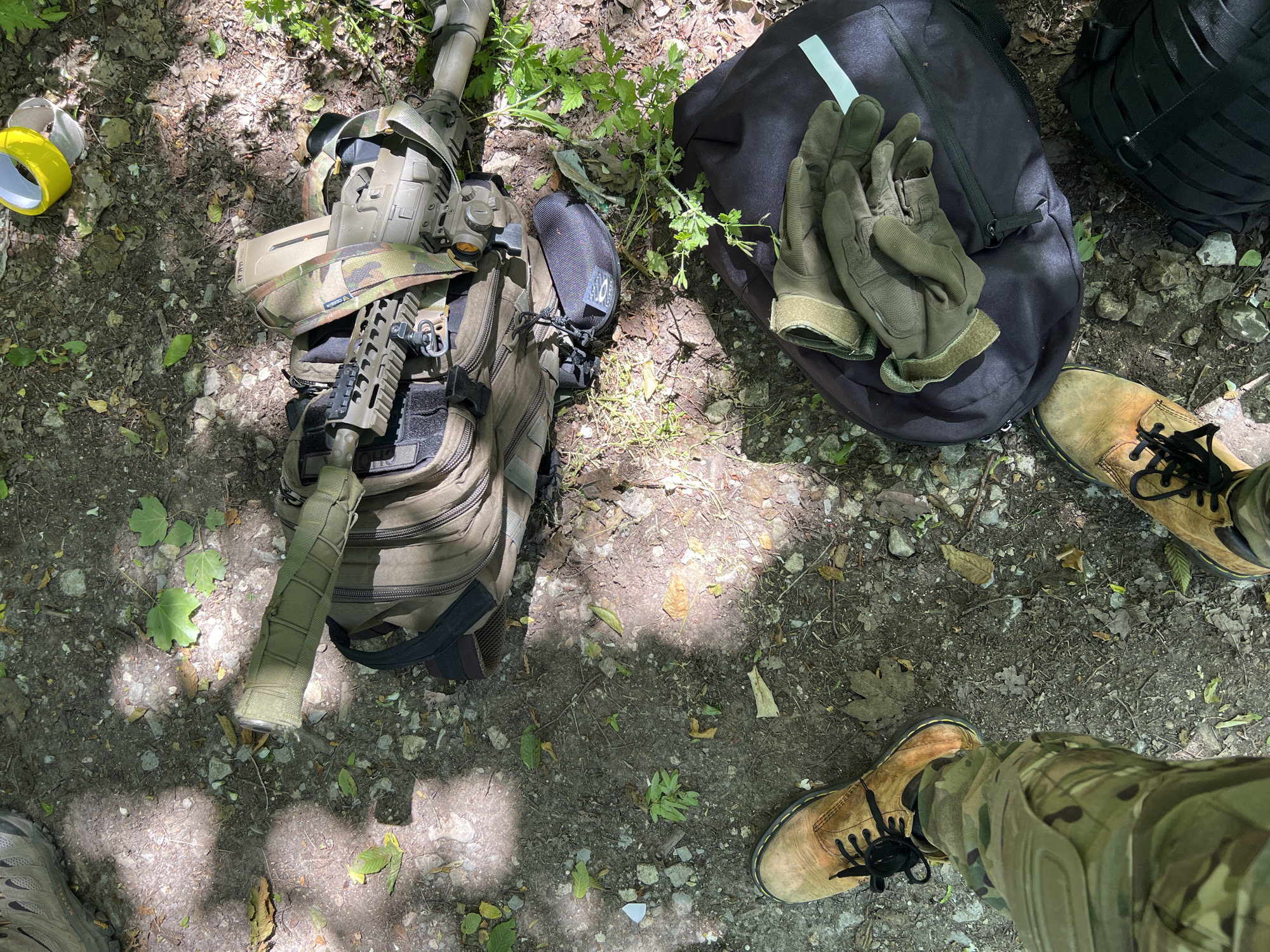 The image shows a ground-level view in an outdoor setting, possibly a wooded area. The ground is covered in dirt, leaves, and small rocks. The scene includes tactical gear: an assault rifle resting on top of a camouflage backpack, a black backpack with a pair of gloves on top of it, and a person's legs wearing camouflage pants and tan boots are visible. A roll of yellow tape is located by the assault rifle. The environment appears to be shaded with dappled sunlight.