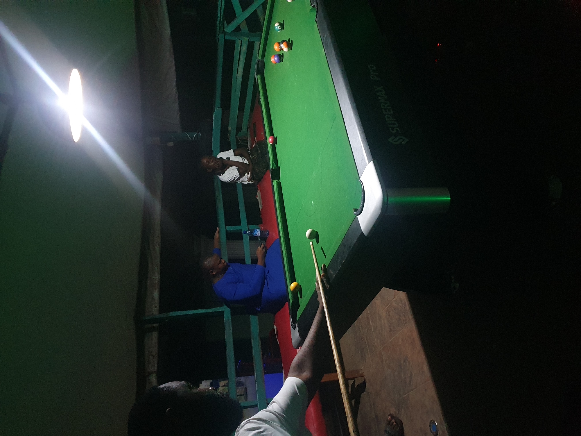 In Lokoja, Nigeria, a group of individuals are gathered around a pool table in an dimly lit outdoor setting. The pool table is green with various colored balls, and a cue stick is being held by one of the men, suggesting a game is in progress. Two men are seated on a bench, one in a blue garment and the other in a white shirt, observing the game. The environment appears to be a casual gathering place, possibly a bar or recreational area, indicated by the presence of a light fixture overhead.