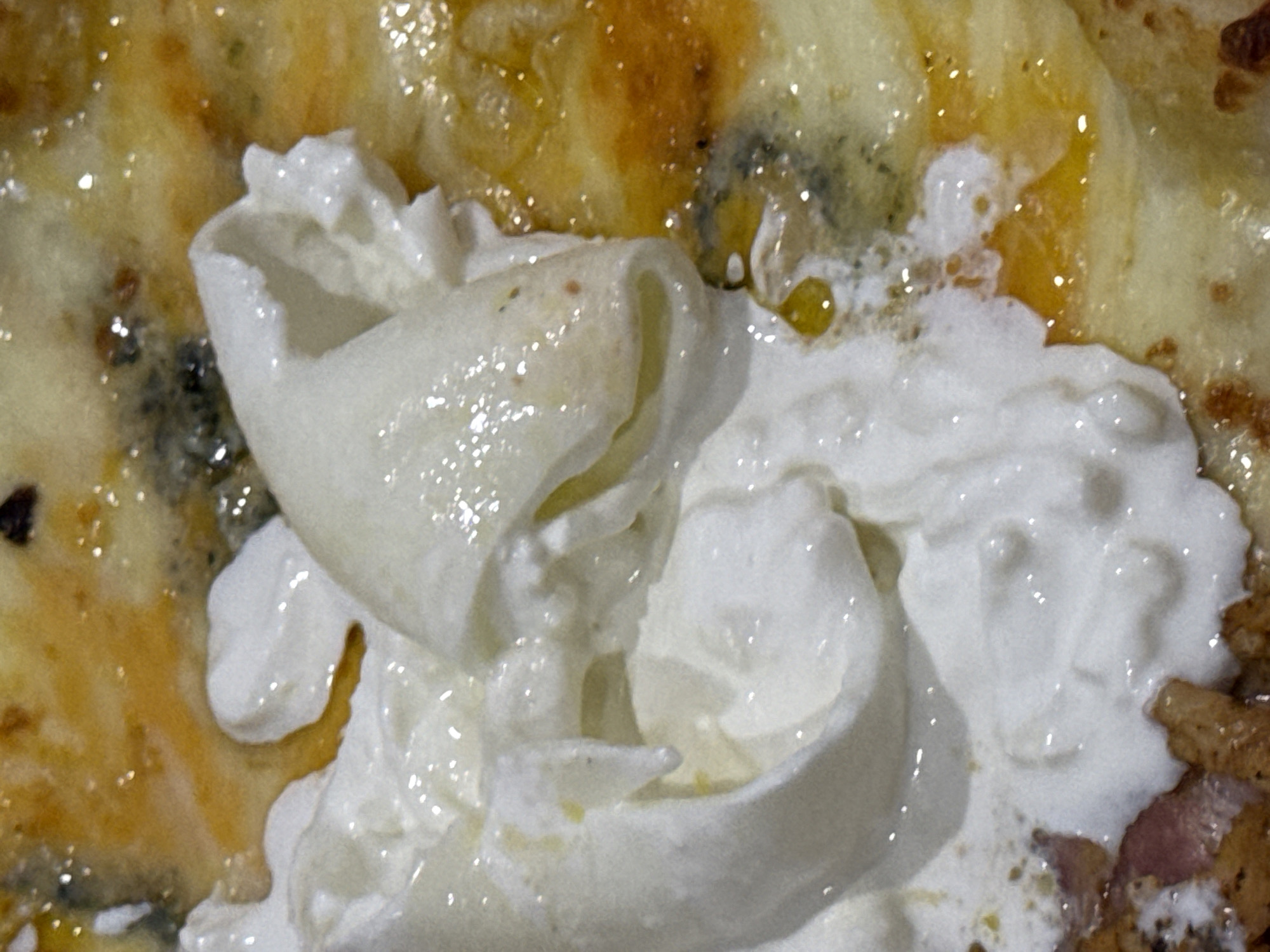 This is a close-up image of food, specifically pizza, with a topping of what appears to be burrata cheese. The cheese is a creamy white, with a slightly glossy texture, and it is layered in a swirled fashion. The crust of the pizza is visible around the cheese, with a golden-brown coloration and some areas that look oily. There is also the appearance of other toppings. No people are visible in the image, and there is no information to determine that the location is in the city of Đà Nẵng, Vietnam.