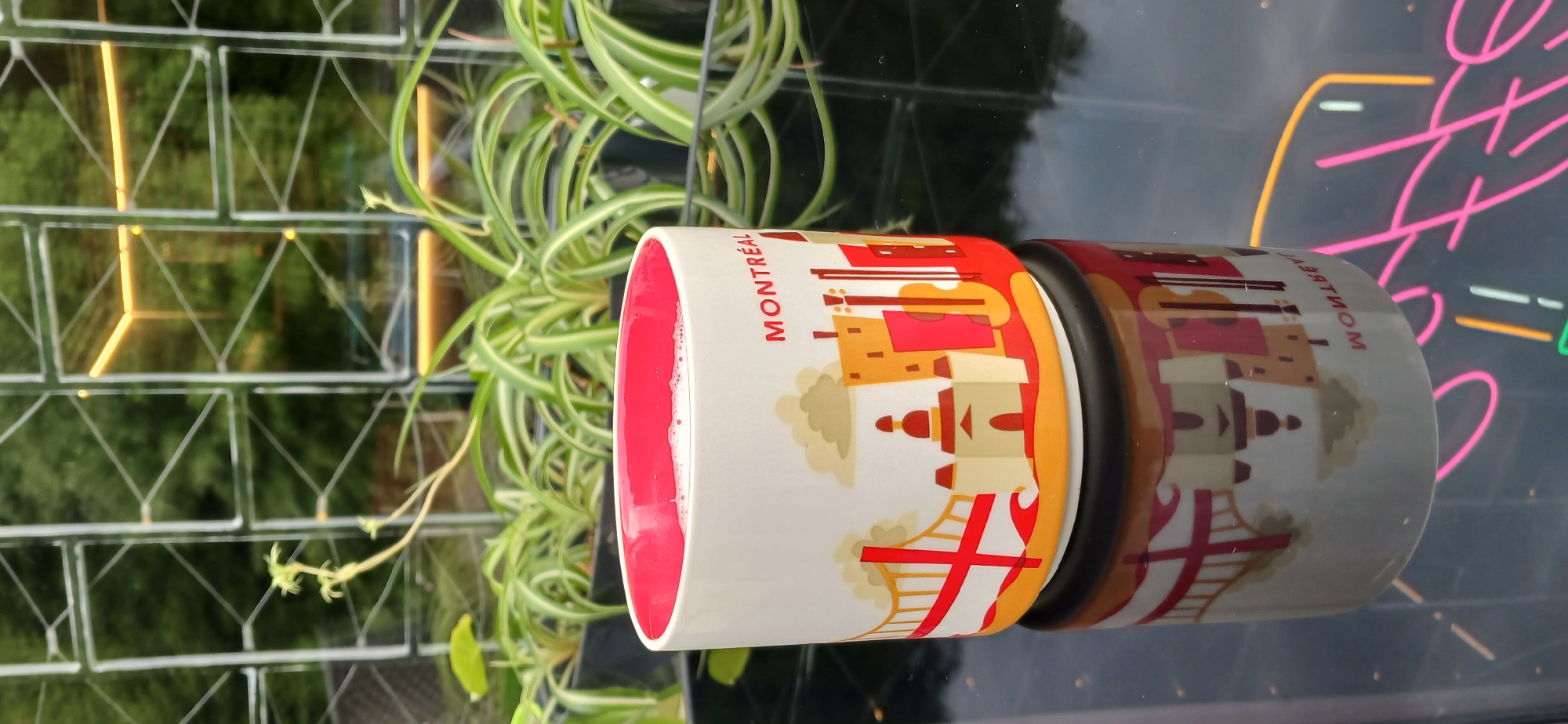 This image features a white mug with a red interior, decorated with a colorful graphic of the Montreal skyline. The mug is filled with a pinkish liquid, likely a beverage. A spider plant with long, variegated leaves is positioned behind and to the left of the mug. The mug sits on a reflective black surface, creating a mirror image of itself and the surrounding environment. In the background, a lattice-like structure with reflective panes is visible, through which a blurred outdoor scene with greenery and warm lighting can be seen. To the right of the mug, a neon sign with pink and yellow lighting is partially visible, reflecting on the surface. The overall scene appears to be indoors, with the window and neon sign suggesting a cafe, shop, or entertainment venue. The lighting suggests it could be evening or a dimly lit indoor space. No people are visible in the image. Text on the mug reads "MONTREAL" in red lettering. The reflection of the mug also shows reversed text that appears to be "LAITMONT" or similar, possibly a souvenir inscription.