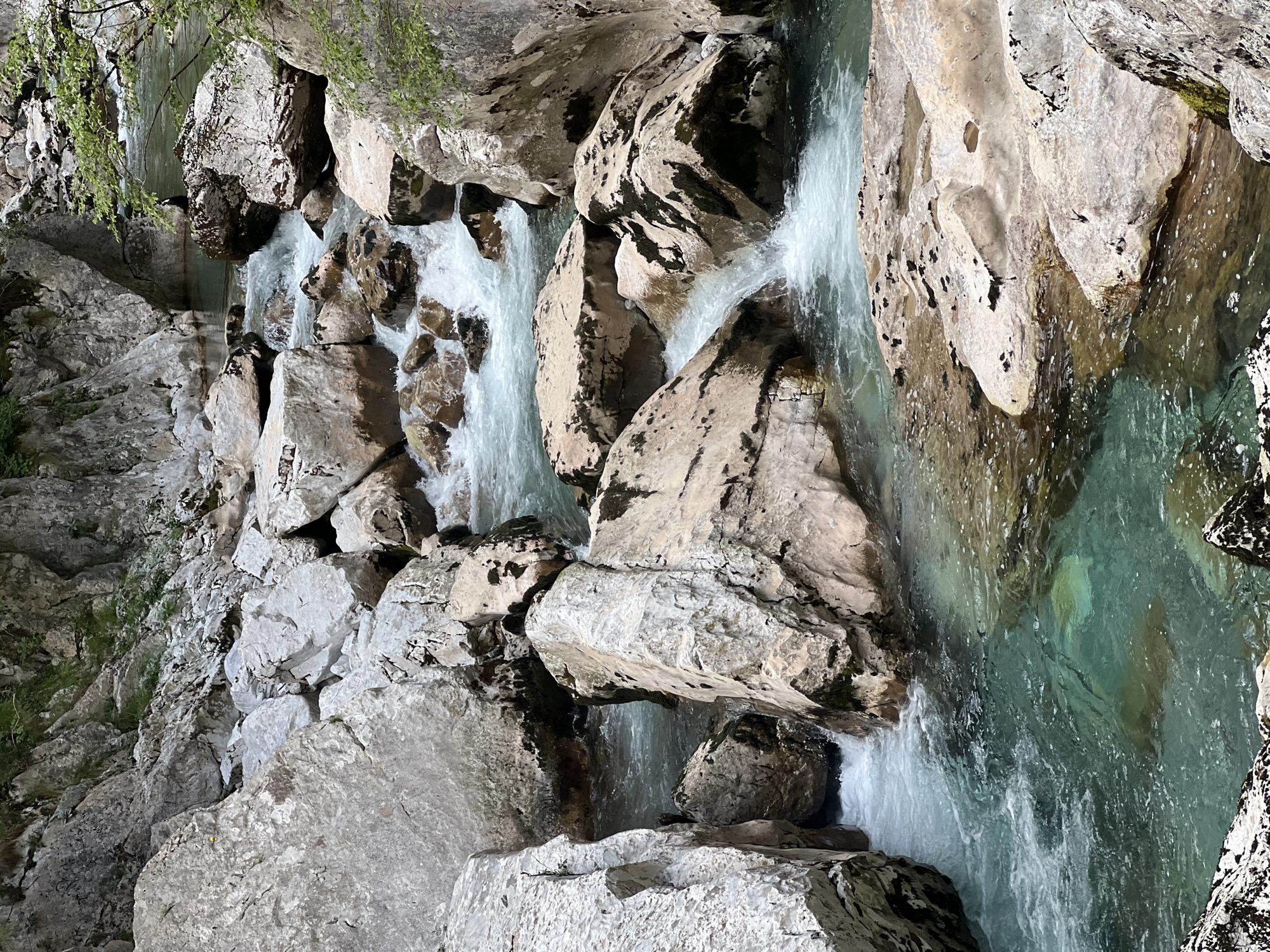 The image depicts a cascading stream flowing through a rocky terrain. The location is presumably near Lazba, Montenegro, evidenced by the geological features characteristic of the region. The stream is characterized by clear, turquoise-tinted water, running over and around a jumble of light-colored, striated rocks. Waterfalls occur at various points as the stream descends. Vegetation is sparsely visible on some of the rocks and along the top edge of the frame. No people, animals, or structures are visible. The overall impression is of a natural, relatively untouched landscape.