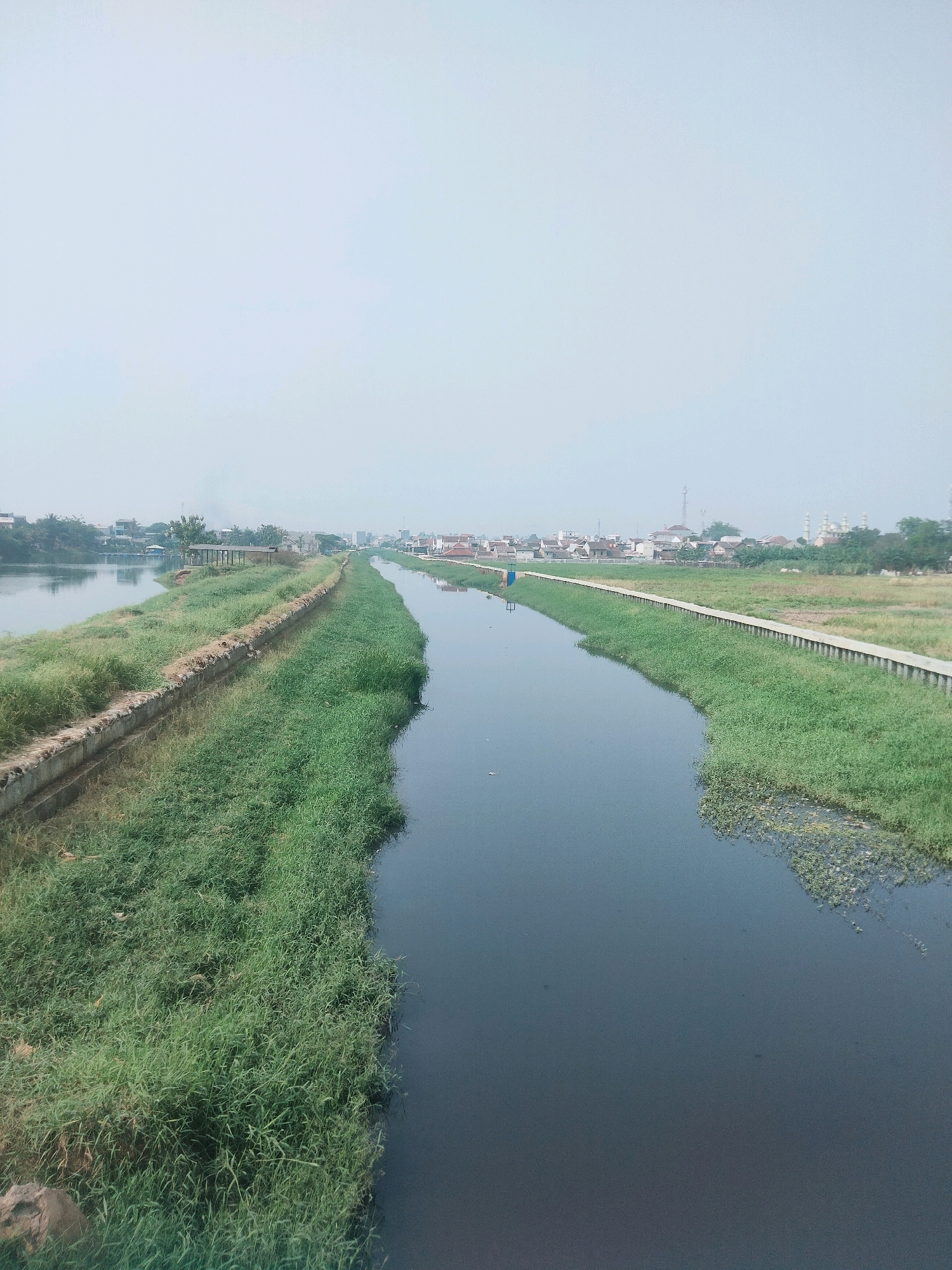 The image shows a canal in Tangerang, Indonesia, with a city skyline in the background. The canal is lined with green grass on both sides. There are buildings and trees visible in the distance. The sky is overcast.