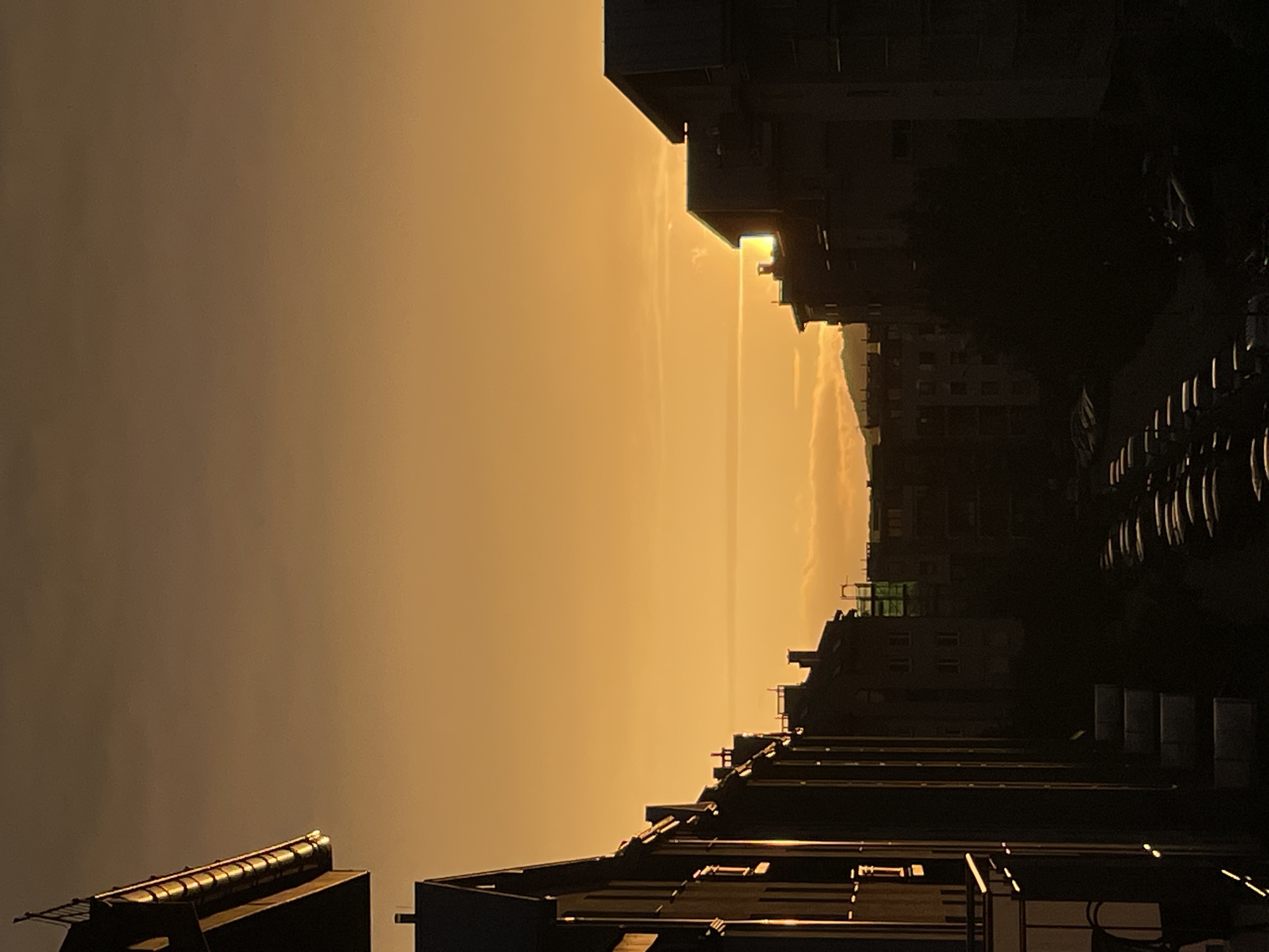 An urban environment is depicted during either sunrise or sunset, characterized by a gradient sky transitioning from dark reddish-brown at the zenith to bright orange-yellow near the horizon. The sun, a brilliant light source, is partially obscured by a multi-story building on the right side of the frame, emitting a horizontal lens flare or light streak across the sky. Wispy clouds are present near the horizon.
On the upper left, a building's roof and gutter system are brightly illuminated by the direct sunlight, highlighting the curved segments of the gutter. A row of dark-silhouetted, multi-story residential buildings extends along the left side of the image, with some windows and balconies catching the sun's reflection as bright vertical and horizontal lines.
On the right, another large building stands in prominent silhouette, with the sun's glare emanating from directly behind or beside its structure. Further into the mid-ground, additional buildings are visible, some with subtle architectural features and one exhibiting what appears to be scaffolding with a green net. Distant mountains or hills are faintly visible as dark outlines on the horizon, particularly in the center-left.
The lower portion of the image is enveloped in deep shadow, revealing a street or alleyway lined with multiple rows of parked cars, whose reflective surfaces are minimally visible. No individuals are discernible within the scene. The location is an unknown city.