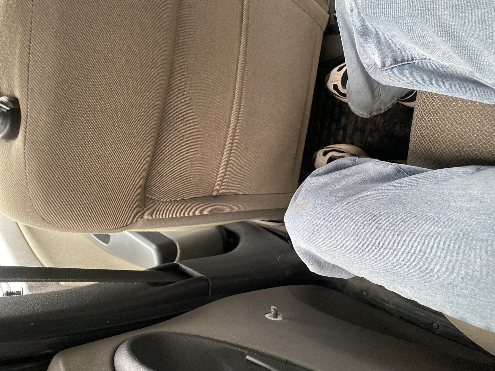 The image shows the interior of a car, specifically the driver's side. A person wearing light blue jeans and white sneakers with black accents is sitting in the driver's seat. The car's seat is light brown fabric. The center console, which is dark gray or black, is visible between the driver's seat and the dashboard. A section of the dashboard, in a lighter gray, is also visible. The floor mat appears to have a patterned texture. The image was taken from a high angle looking down and to the side. There is no visible information that can be used to identify the location in Chaikovsky, Russia.