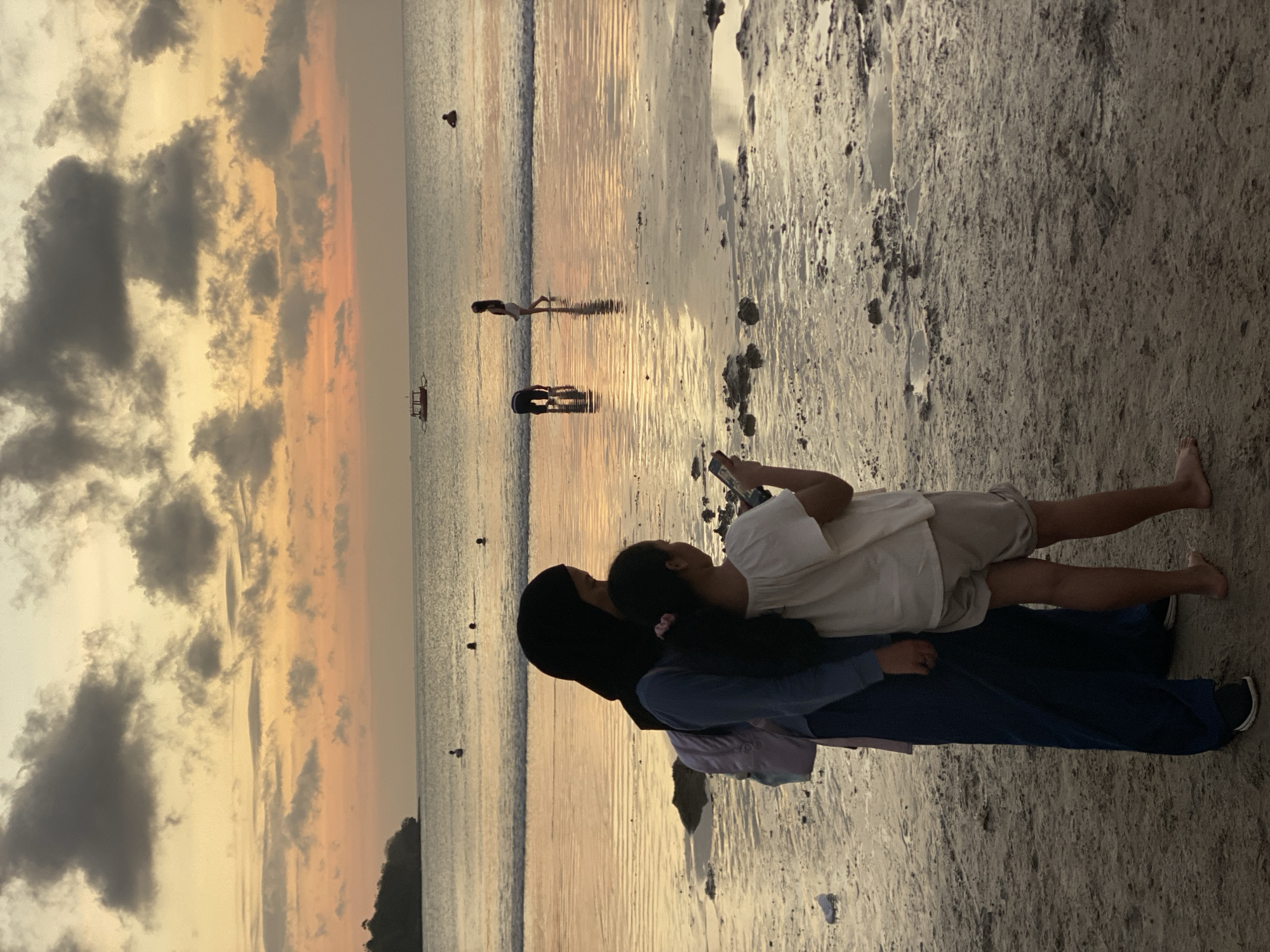 This is a vertical photograph taken at a beach during sunset. The foreground is dominated by a sandy shore with scattered rocks and some tide pools reflecting the sky. Two people are the main subjects in the foreground, positioned vertically in the frame. The person closer to the viewer is wearing a dark hijab and a blue garment, with a lighter purple garment visible underneath. They are embracing a person in front of them, who is wearing a white top and light-colored shorts. The person in front is looking down at a smartphone they are holding. Their legs and feet are clearly visible, with the person in the blue garment also showing their feet.
In the middle ground, the calm sea stretches out, its surface shimmering with the reflection of the sunset. Several small boats are visible on the water, and a few distant figures can be seen walking or standing in the shallow water. The water's edge creates a distinct line between the wet sand and the open sea.
The background is characterized by a dramatic sunset sky. Dark, textured clouds are scattered across the sky, interspersed with areas of soft orange and yellow hues, indicating the sun is low on the horizon. The overall atmosphere is serene and peaceful, suggesting a relaxed evening at the beach. The time of day is clearly sunset, and the weather appears calm. The location is identified as Kuta Selatan, Indonesia, a known coastal area. There is no visible text in the image.