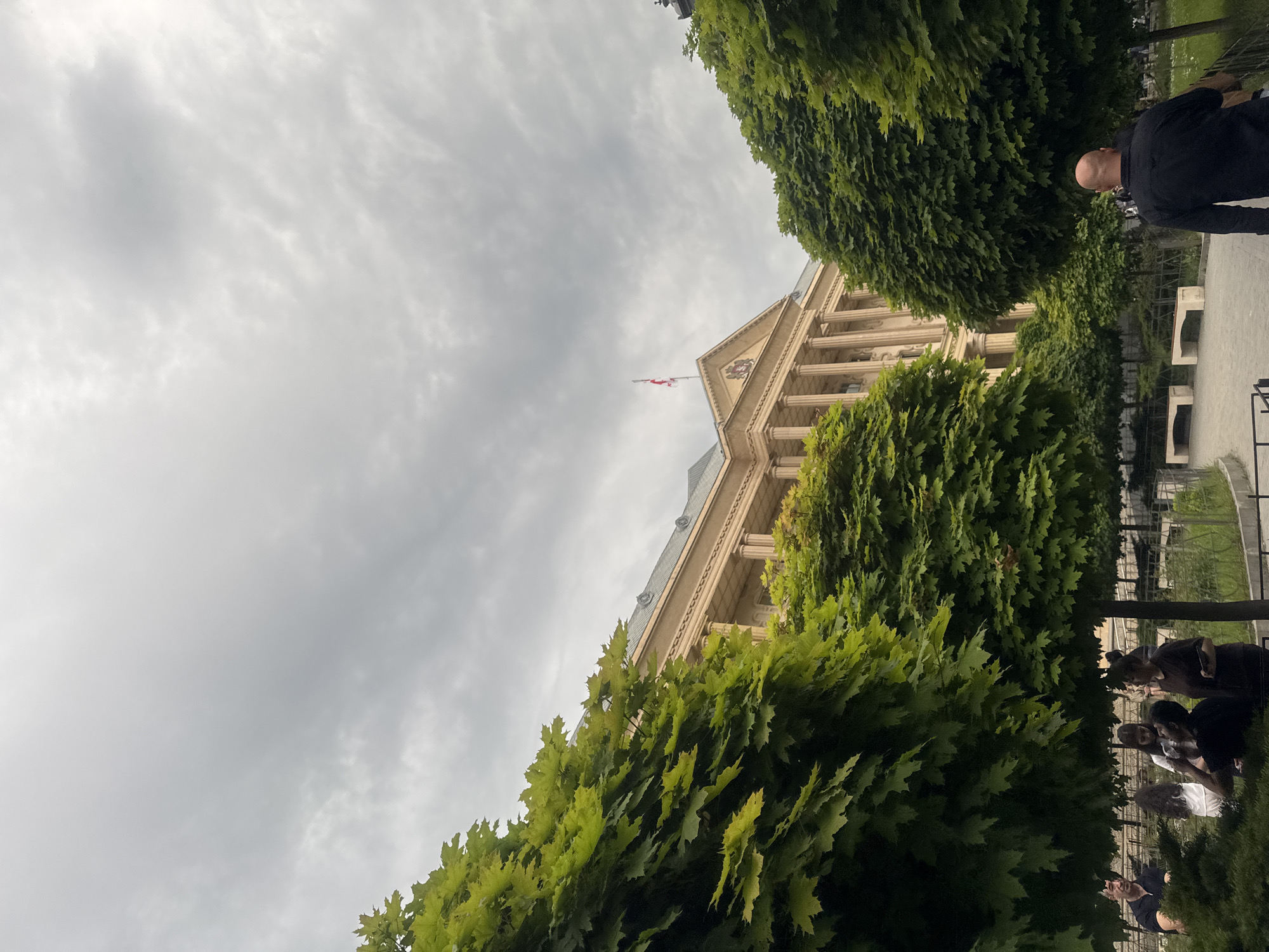 The image shows the Parliament of Georgia in Tbilisi. The building has a classical style with columns and a flag on top. The sky is cloudy. Green trees frame the building. A few people are visible in the lower right and upper right areas of the image, engaging in casual activities.