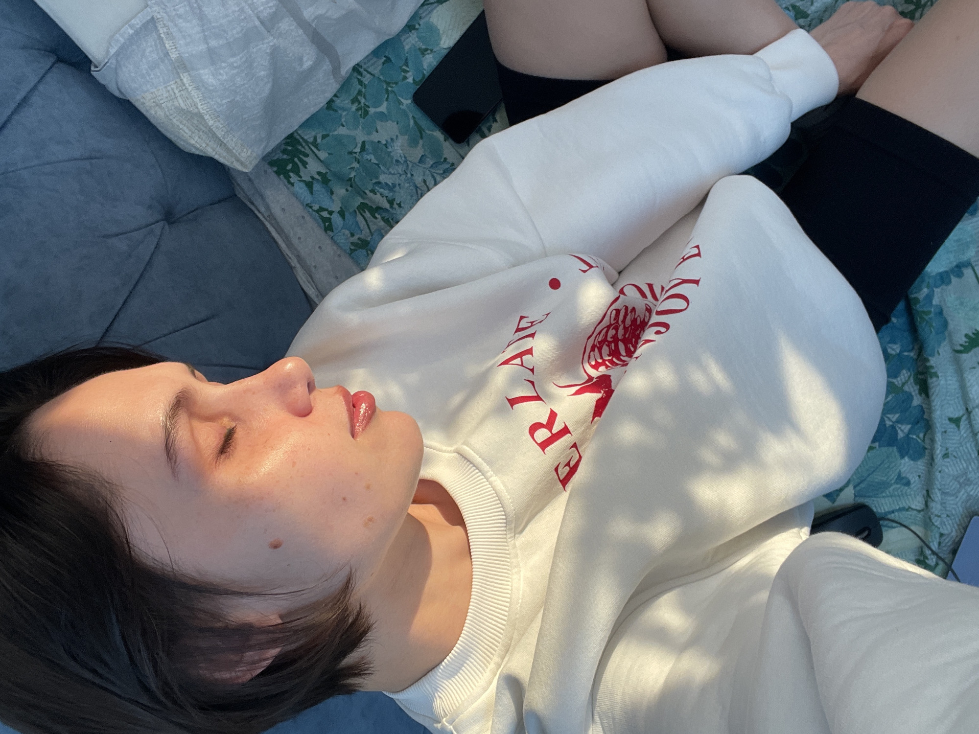 A fair-skinned person with short, dark hair lies on their back, eyes closed, on a blue-gray tufted cushion, wearing a white sweatshirt with a red floral and lettering design. The person's face is illuminated by sunlight, highlighting their features. Adjacent to the cushion, there's a patterned floral blanket in shades of blue, green, and white. On the blanket, partially obscured by the sweatshirt, lies a dark rectangular object, possibly a phone. The person's upper legs are visible, clad in black shorts. A portion of another patterned blanket appears in the background. Location is unidentifiable as Izhevsk, Russia based on visual elements.