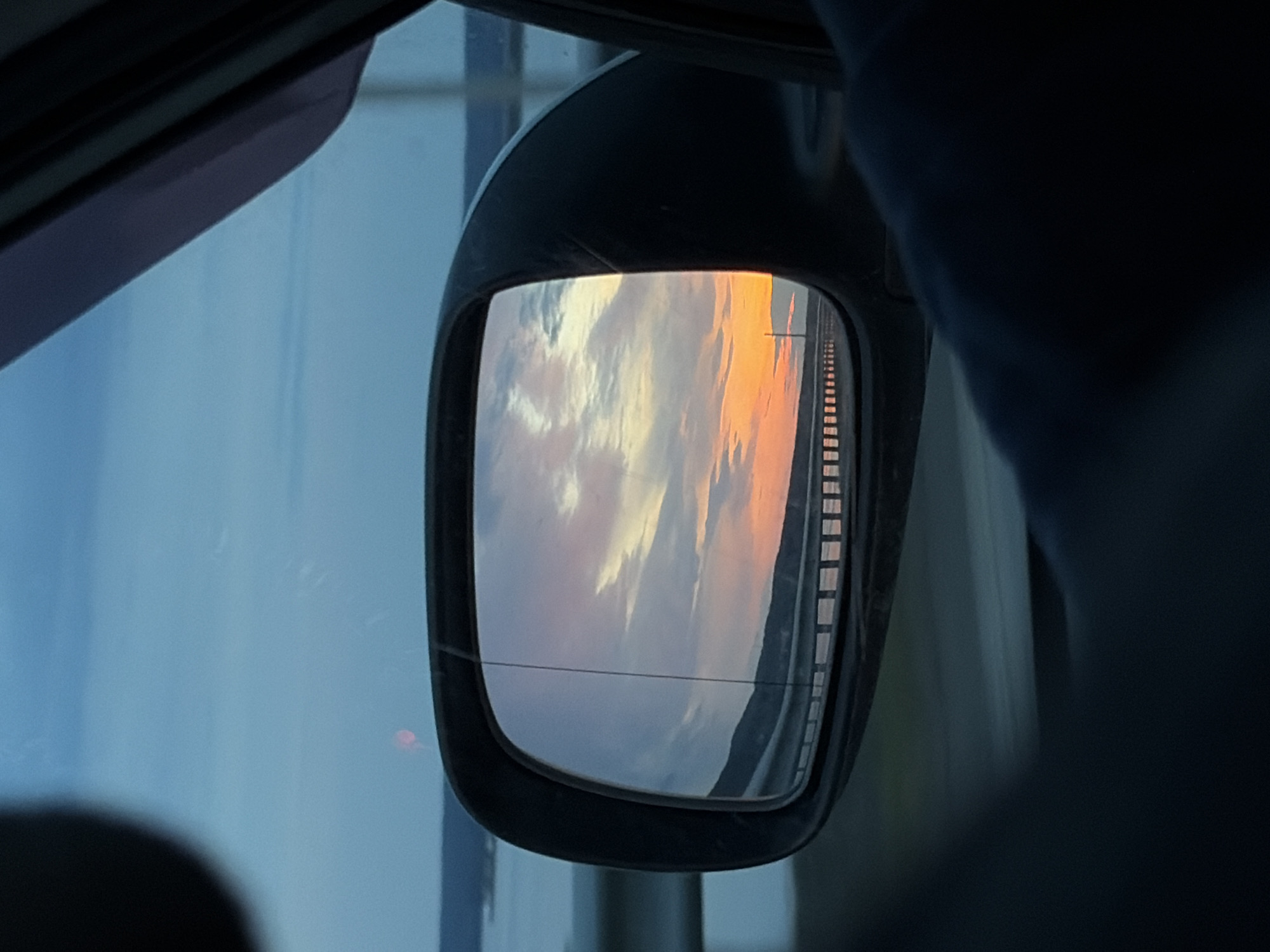 The image shows a vehicle's side mirror reflecting a sunset with pink and orange clouds over a highway or bridge. The mirror's black frame is visible, along with parts of the vehicle's interior, like the window and possibly a seat. There are no people directly visible. No specific details in the image indicate its location is Chaikovsky, Russia.