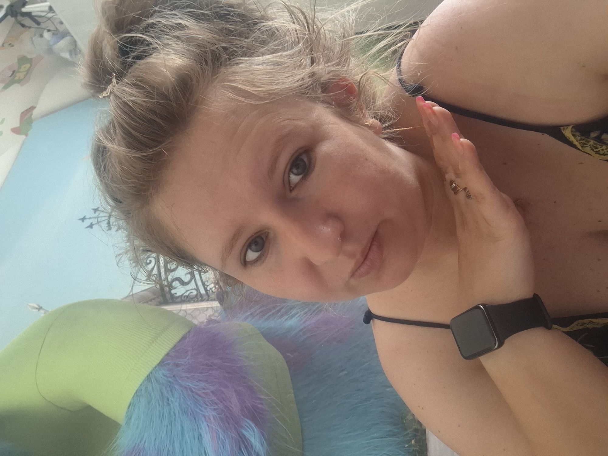 A woman with blonde hair pulled up in a messy bun takes a selfie in front of a blurred background. She is wearing a black spaghetti strap top and a smartwatch on her left wrist. Her right hand is resting on her neck, and her left hand is on her cheek. The background appears to be a room with blue and light green decor, possibly a child's room, with a large stuffed toy on the left side of the frame. The woman has light skin and blue eyes. The overall lighting suggests it is daytime. There are no specific details in the image that indicate the location is in Shymkent, Kazakhstan.
