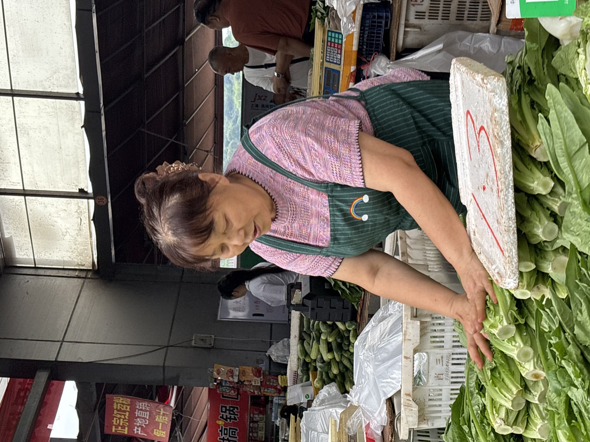 The image shows a woman in Jiangjin, China, working at a produce stand in an open-air market. She wears a pink and white patterned shirt and a green striped apron with a rainbow on it, and her gray hair is pulled back. She is reaching into a bin of fresh, leafy green vegetables, likely sorting or arranging them. In the background, other vendors and customers are visible. The location appears to be a traditional, local market, with hanging banners and signs in Chinese characters. The bins of produce are arranged on crates. A sign with red markings stands amid the vegetables. Further back, there is also a pile of green cucumbers. The market setting is underneath a shelter.