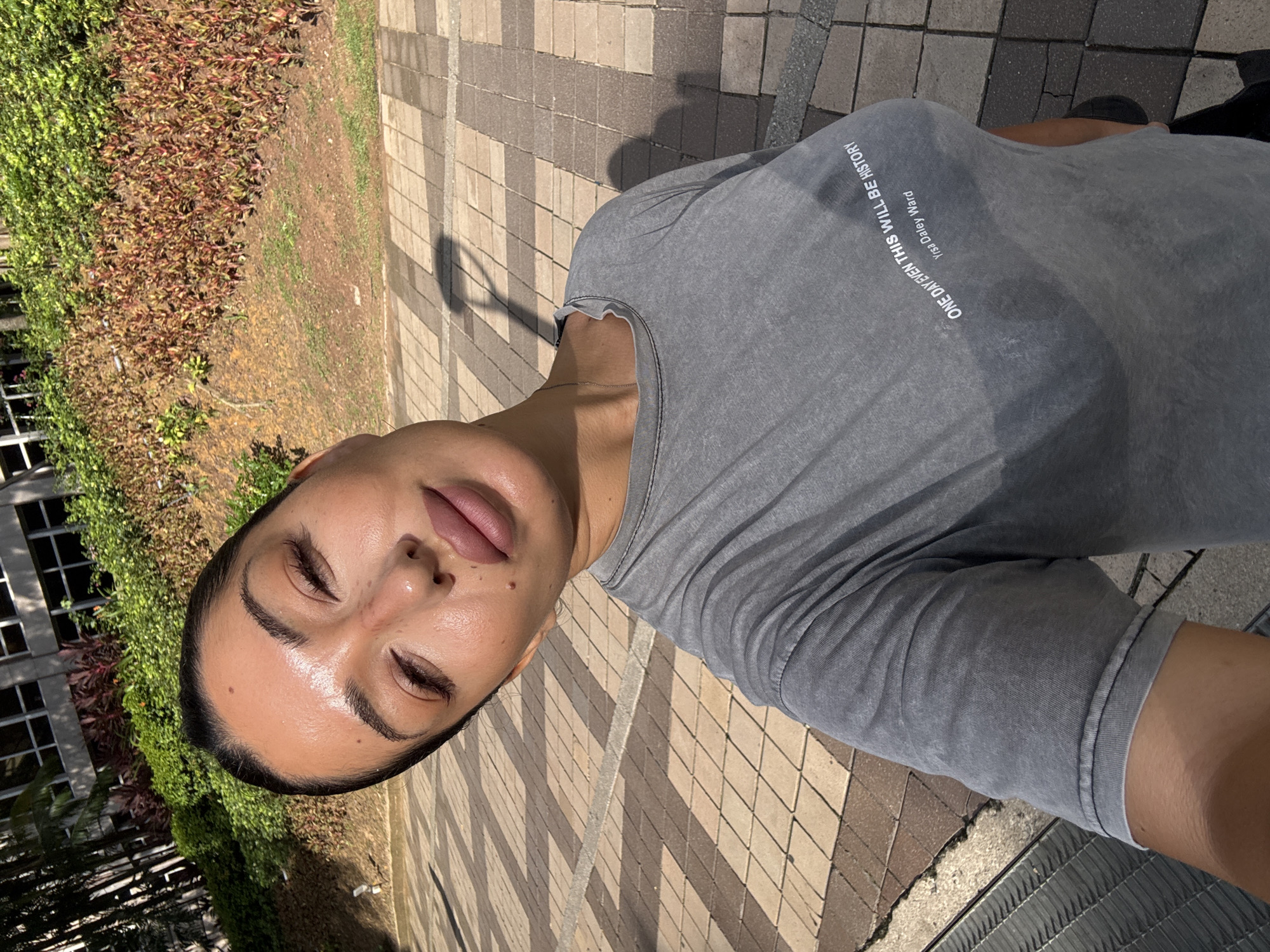 A selfie taken outdoors on a paved area. The subject is a woman with her eyes closed, wearing a grey t-shirt with the text "ONE DAY EVEN THIS WILL BE HISTORY Yisa Daley Ward." The background includes green foliage, a building with windows, and a patterned brick walkway. The photo appears to be taken in bright daylight, casting shadows across the scene. It is unknown if this photo was taken in Kuala Lumpur, Malaysia.