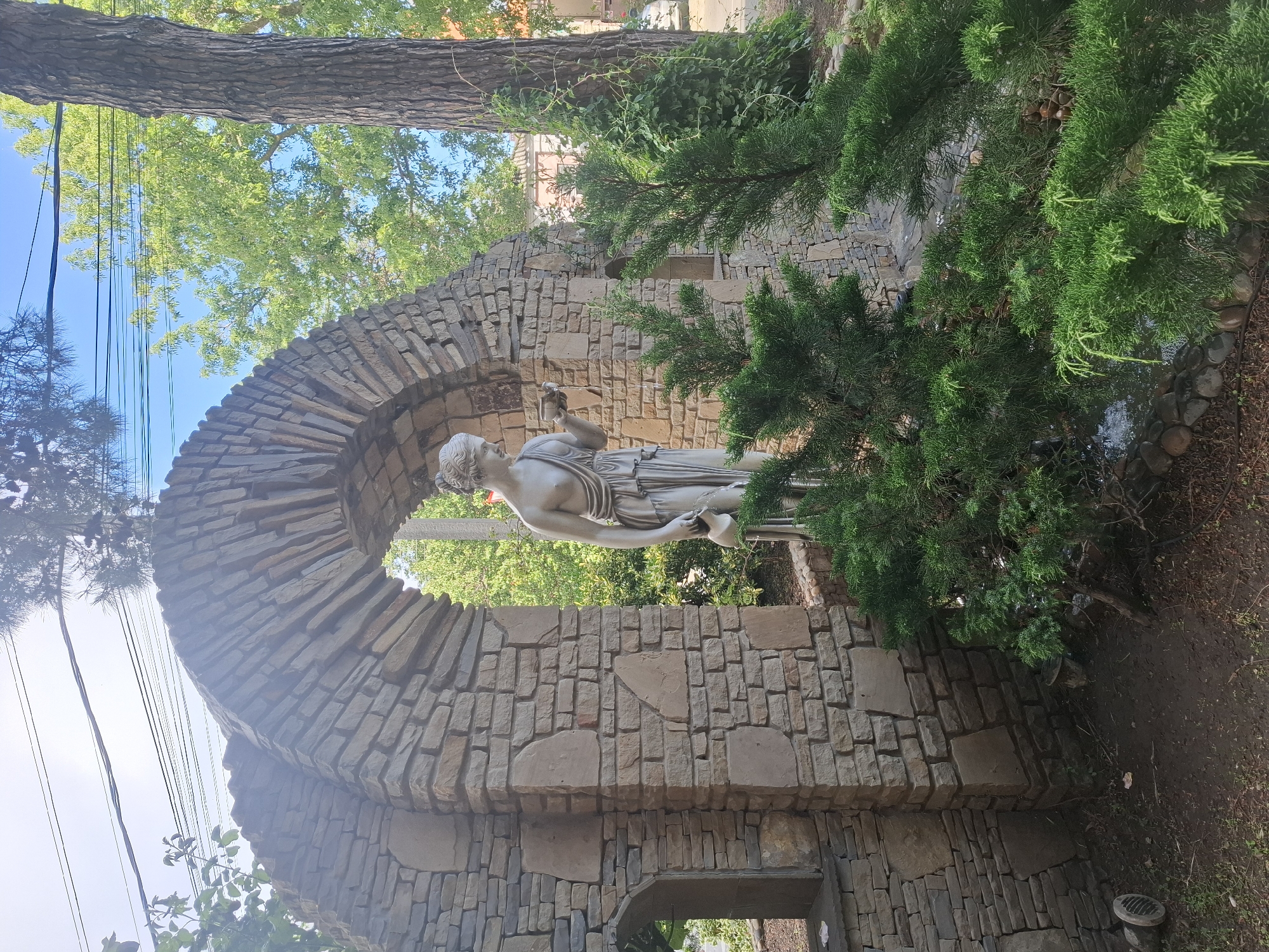The photograph captures a sunlit outdoor scene in Anapa, Russia, featuring a stone archway with a statue of a woman holding a pitcher. The statue, rendered in a classical style, stands within the archway, which is constructed from rough-hewn, light-colored stones. Behind the arch, lush green trees and foliage are visible, suggesting a park or garden setting. The sky above is a bright blue with some wispy clouds. In the foreground, dark green coniferous shrubs partially obscure the base of the arch and the statue. The ground appears to be a mixture of dirt and small stones. The overall impression is one of a tranquil, decorative garden feature.