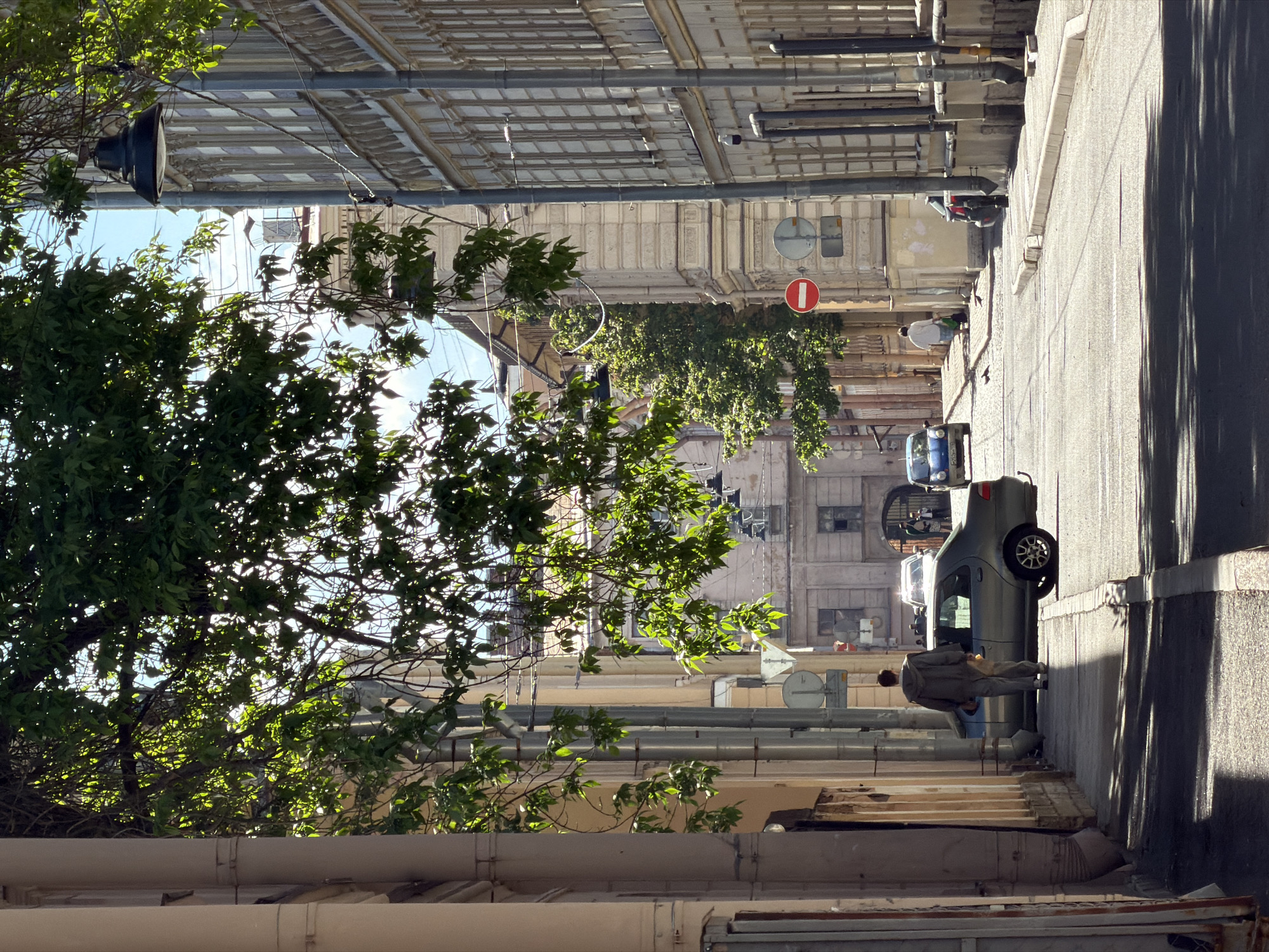 The image depicts a sunlit urban street in Saint Petersburg, Russia, looking into the distance. Lush green trees with bright foliage line the left side, with strong sunlight filtering through their leaves, creating dappled patterns. The street is flanked by multi-story buildings of classic European architectural style, featuring multiple windows and external pipes.
On the right side of the street, a paved sidewalk runs alongside the buildings. Distinct, elongated shadows are cast across the street and sidewalk, indicating bright sunshine, likely in the morning or late afternoon. A gray hatchback car is parked on the right, and a person dressed in a light-colored jacket and pants stands beside its passenger door, appearing to hold a light-colored bag. Further down the street, a blue car is visible, and beyond that, a red moped or scooter is parked.
Several street signs are present, including a prominent red circular "no entry" sign suspended above the street, and a circular blue sign with a white horizontal stripe adjacent to it. Above the buildings and through the tree canopy, various wires, cables, and rooftops or overhead structures are visible against a bright, clear sky. The scene captures a quiet moment on a typical city street.