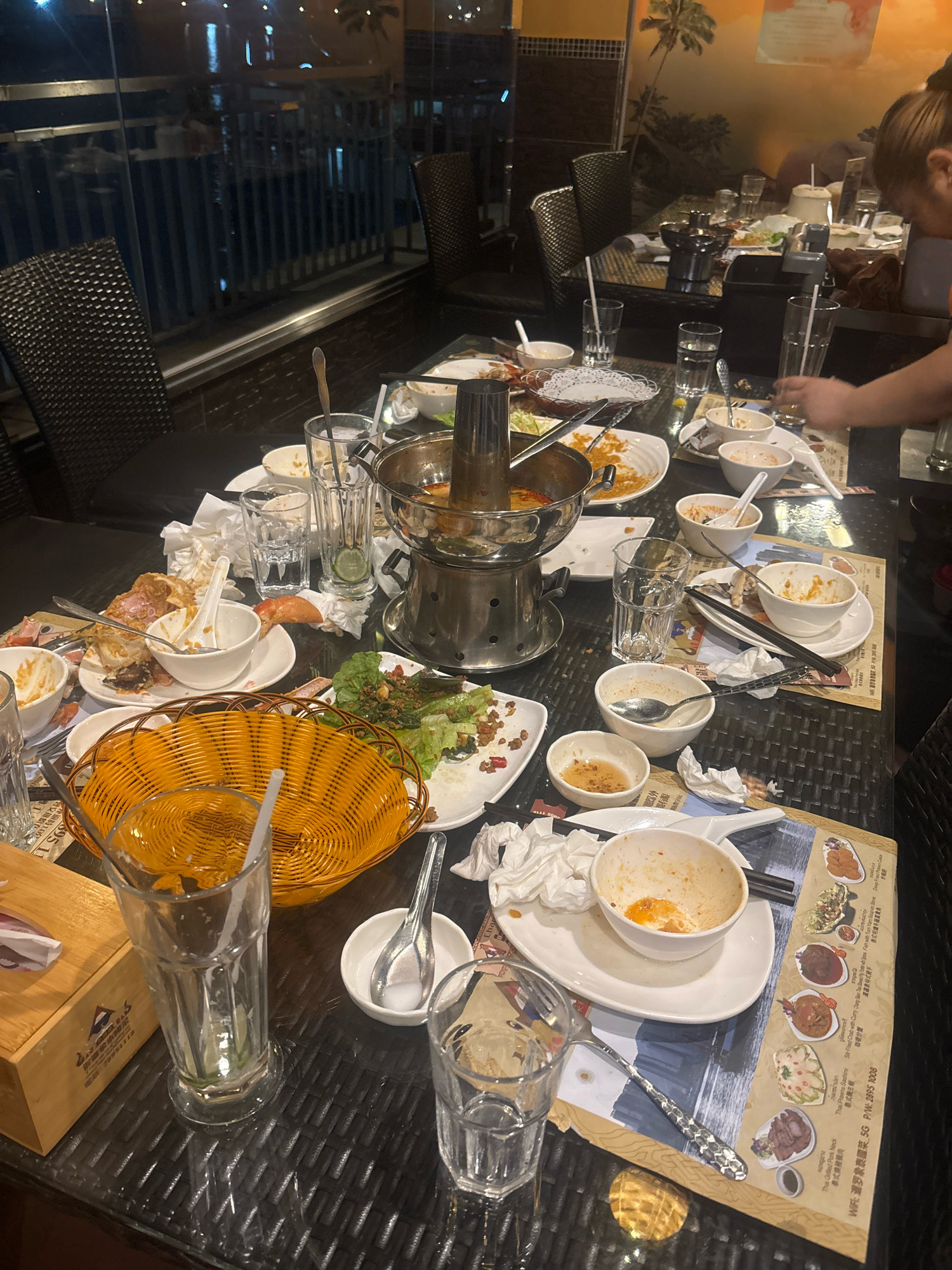 The image depicts a messy table in a restaurant with a view of what appears to be a waterfront area with buildings and lights, possibly in Macau, China. The table is laden with used dishes, cutlery, and crumpled napkins, indicating the aftermath of a meal. A large metal pot, likely a hot pot, is visible in the center, surrounded by empty bowls, plates with remnants of food, and glasses. A woven orange basket is placed on the table, and a wooden tissue box is visible in the foreground. The background shows chairs and a wall decorated with a mural of palm trees. A person's arm is partially visible on the right side of the image, suggesting someone is clearing or interacting with the table.