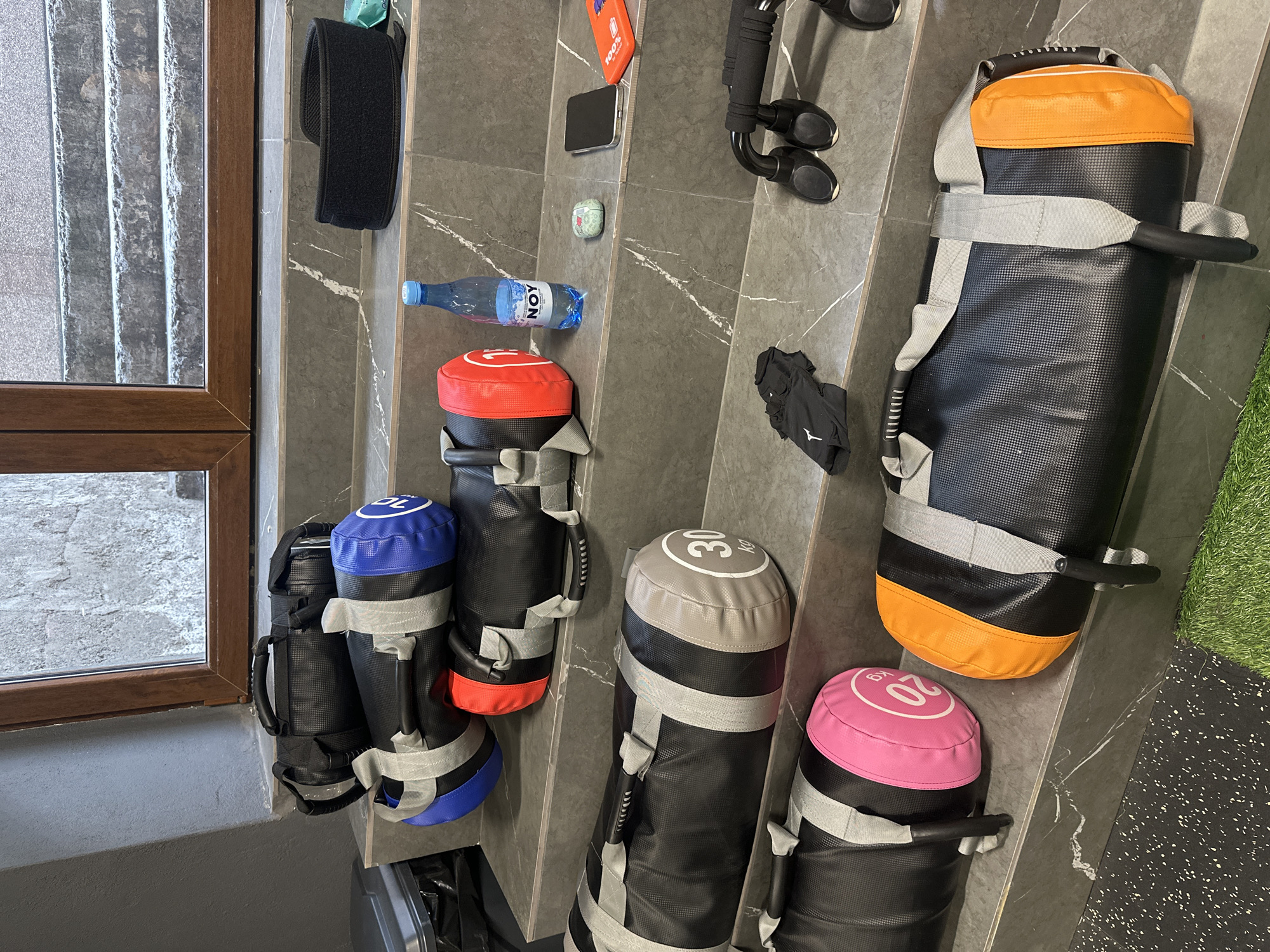 A low-angle medium shot of a gym wall shows several weight training bags of various weights. From left to right, there is a bag with a blue end, then a bag with a red end, then a bag with a gray end, then a bag with a pink end, and finally a bag with an orange end. Other items on the wall include a black velcro strap, a clear water bottle, a black cell phone, a pair of white AirPods, a pair of black gloves, and a piece of gym equipment. A dark wooden window frame is on the left side of the frame. The wall is dark gray with white streaks. The location is Yerevan, Armenia.