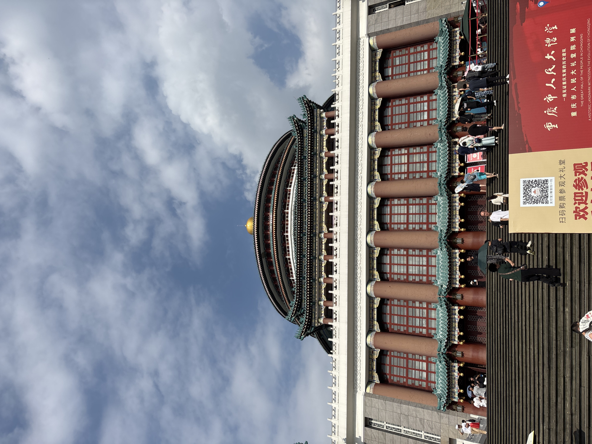 The image shows a low-angle, eye-level shot of the Great Hall of the People in Chongqing, Yuzhong District, China, against a partly cloudy blue sky. The building features a distinctive multi-tiered, circular roof with a golden dome at its apex. The facade is adorned with traditional Chinese architectural elements, including intricate green tiled roofs, ornate eaves, and rows of large, red-framed windows. The building is supported by massive, reddish-brown cylindrical columns. A grand, wide staircase leads up to the building's entrance. Several people are visible on the stairs, some ascending, others standing or walking. One person in the foreground is holding a green umbrella. A large red banner with Chinese characters and a QR code is positioned at the bottom right of the image, indicating an exhibition. The overall scene conveys a sense of grandeur and cultural significance.