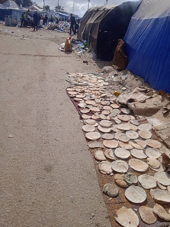 The image depicts an outdoor scene in Khan Yunis, Palestinian Territory, characterized by makeshift shelters and widespread refuse. In the foreground, a woven mat is laid on asphalt, displaying dozens of round, flatbread-like items varying in color from light beige to darker tan, some appearing irregular in shape or discolored. Adjacent to the mat, against a dirt embankment, lies an accumulation of trash, including plastic and cardboard. Behind this, two makeshift shelters are visible: a dark tarpaulin structure from which smoke or steam emanates, and a bright blue tarpaulin structure. In the midground, a dirt road leads into the distance. The background reveals an extensive area populated by numerous makeshift tents and shelters constructed from various materials, including white and dark fabrics. Several individuals are present among these structures and substantial piles of garbage; one person is seen bending over near a trash pile, while others walk or stand. The sky is partly cloudy, and utility poles with overhead lines are visible, along with distant palm trees.