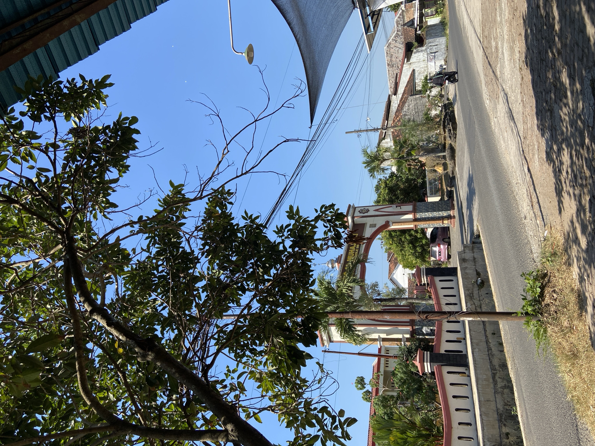 The image depicts a street scene in Margorejo, Indonesia, on a clear, sunny day. In the foreground, a tree with lush green leaves partially obstructs the view of a large arched gateway. The gateway is ornate, with white and red detailing, and appears to be an entrance to a building or institution, with text visible above the arch. A white wall with red trim and vertical black openings runs along the side of the road.
Beyond the gateway, a pink car is parked. The road itself is asphalt, and to the right, there are several houses with tiled roofs. A person on a motorcycle is riding down the road, moving away from the viewer. Overhead, electrical wires crisscross the sky, and a street lamp is visible. A large, dark fabric shade is stretched over a structure on the right side of the image. The overall impression is a typical street view in a tropical urban or semi-urban environment.