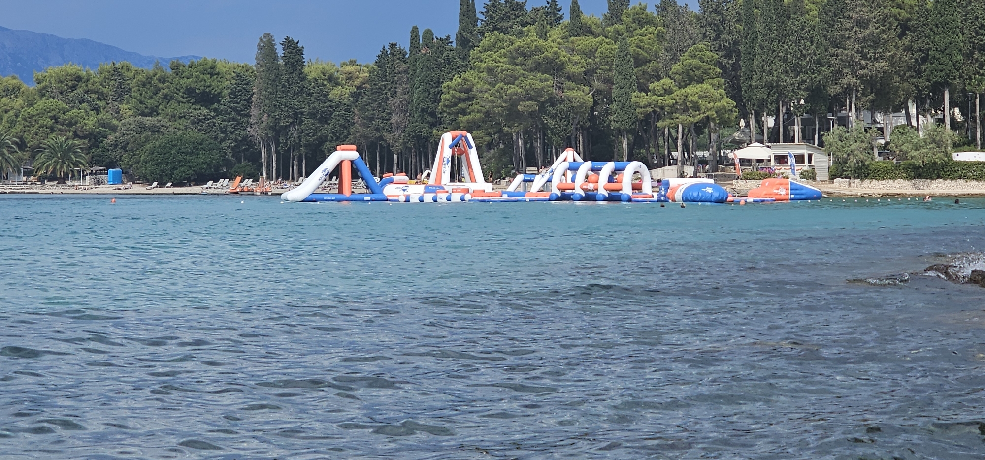 An aerial, panoramic view captures a vibrant inflatable water park situated on the turquoise waters of the Adriatic Sea, adjacent to the shoreline of Supetar, Croatia. The inflatable structures, a combination of blue and orange, feature slides, climbing walls, and arched obstacles, inviting aquatic recreation. In the background, a lush green coastline is dotted with cypress trees and pine trees, providing a striking contrast to the clear blue water. The beach area is furnished with lounge chairs and umbrellas, suggesting a leisurely atmosphere. Several individuals are visible in the water, some interacting with the inflatable park and others swimming. A small, white building with a red roof and a cafe umbrella are also present along the beachfront. The sky above is a mixture of blue and grey clouds, with distant mountains visible on the horizon. The overall scene depicts a popular tourist destination offering a blend of adventure and relaxation.