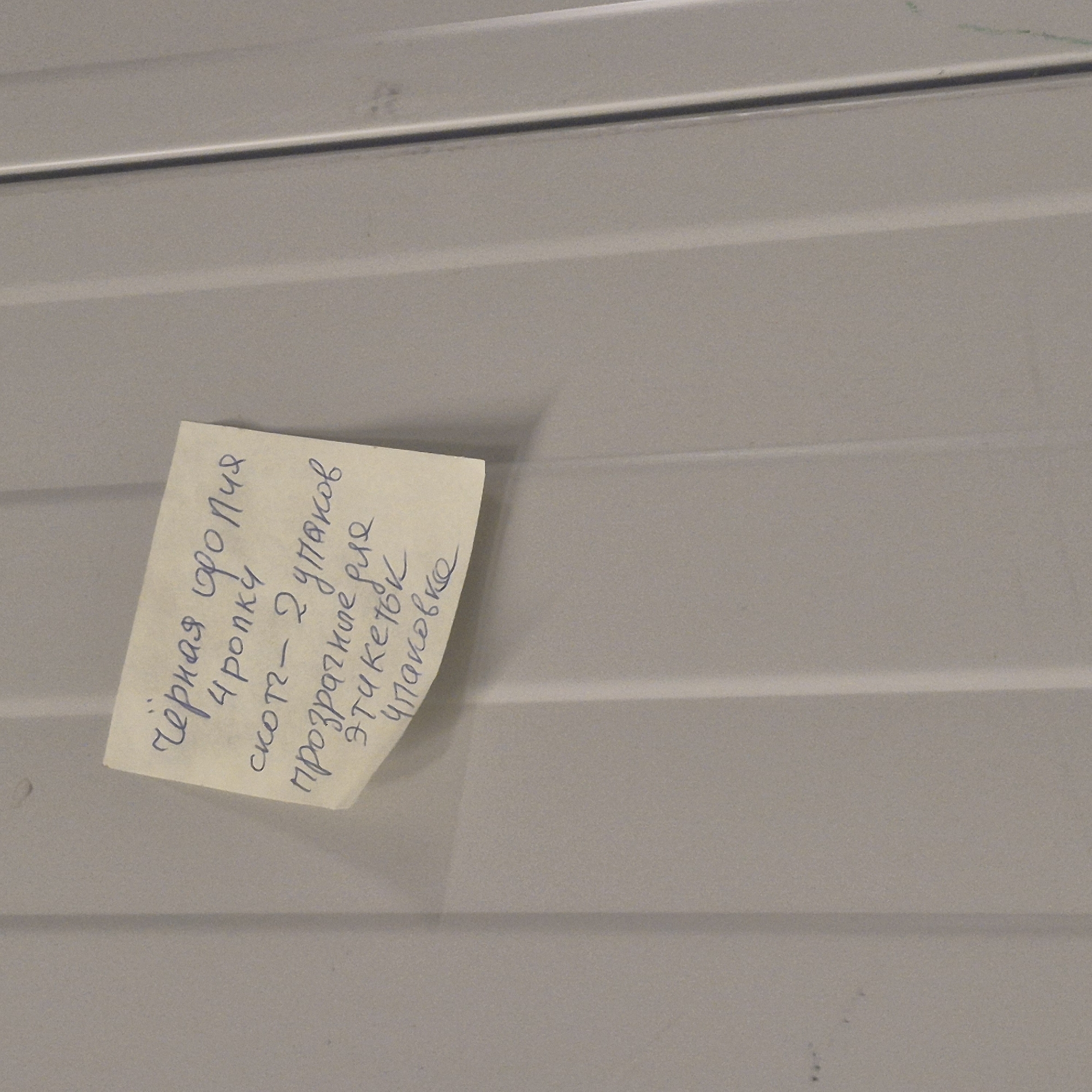 A small, yellow sticky note with handwritten text in Russian is attached to a light gray, ribbed surface. The note is angled and partially obscures the ribbed texture of the surface, which appears to be made of metal or plastic. The lighting is even, suggesting an indoor environment. The text on the note is written in blue ink.
The text reads:
"чёрная фолия
4 ропкц
скотч- 2 упако
прозрачные для
этикеток
упаковко"
This translates to:
"black foil
4 rolls
scotch tape - 2 packs
transparent for
labels
packaging"
The image captures a close-up of the sticky note and the surface it's attached to, with no people or other significant objects visible. The scene is static and focused on the handwritten note, suggesting it's a list or reminder of items. The location context of Nowa Wieś Książęca, Poland, does not provide additional clues within the image itself.